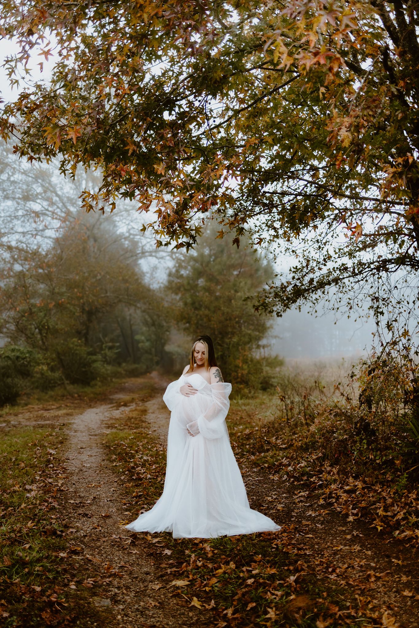 Pregnant woman in an elegant white gown posing on a foggy autumn forest path under colorful trees.