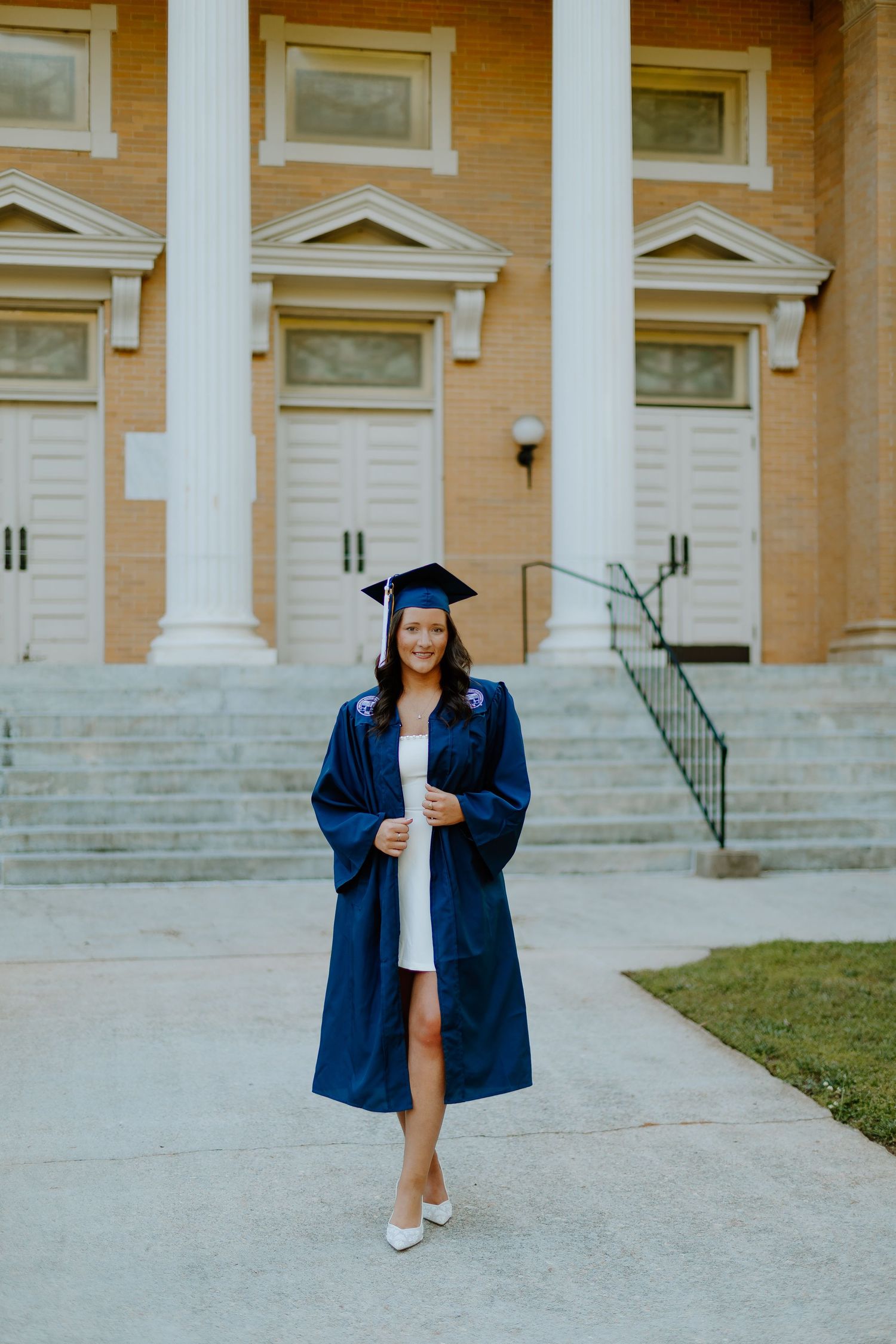 Young woman in navy graduation cap and gown smiling in front of a brick university building.