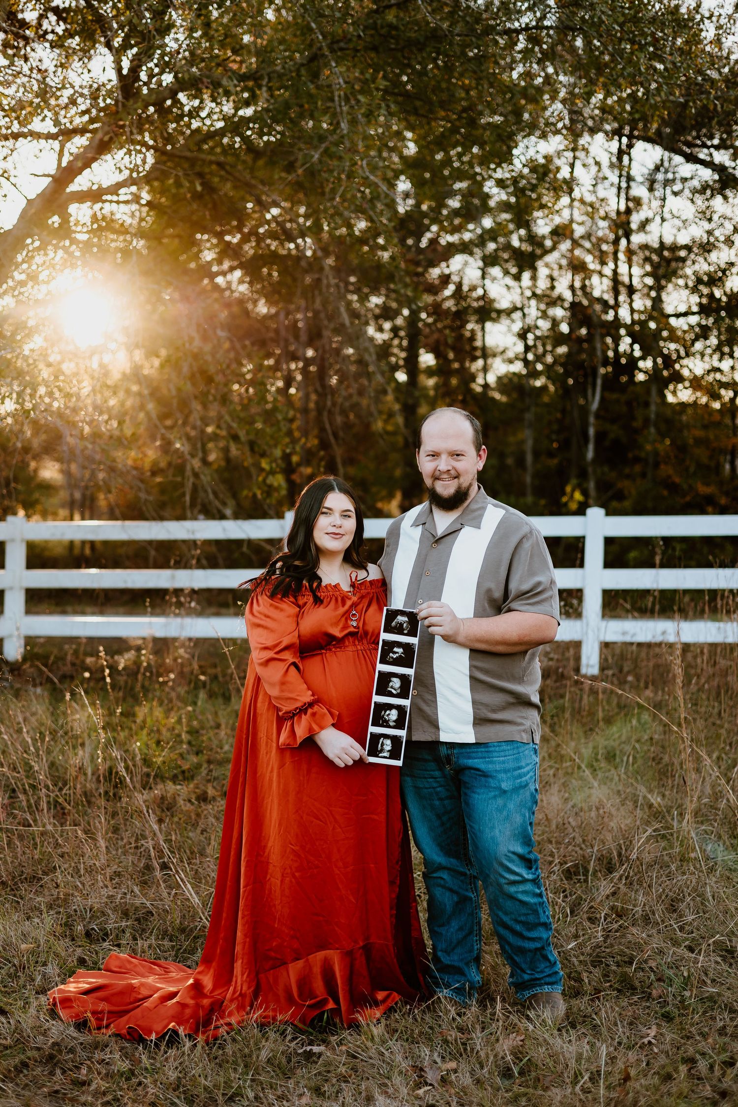 Expecting couple holding ultrasound photos by a white fence in a field at golden hour sunset.