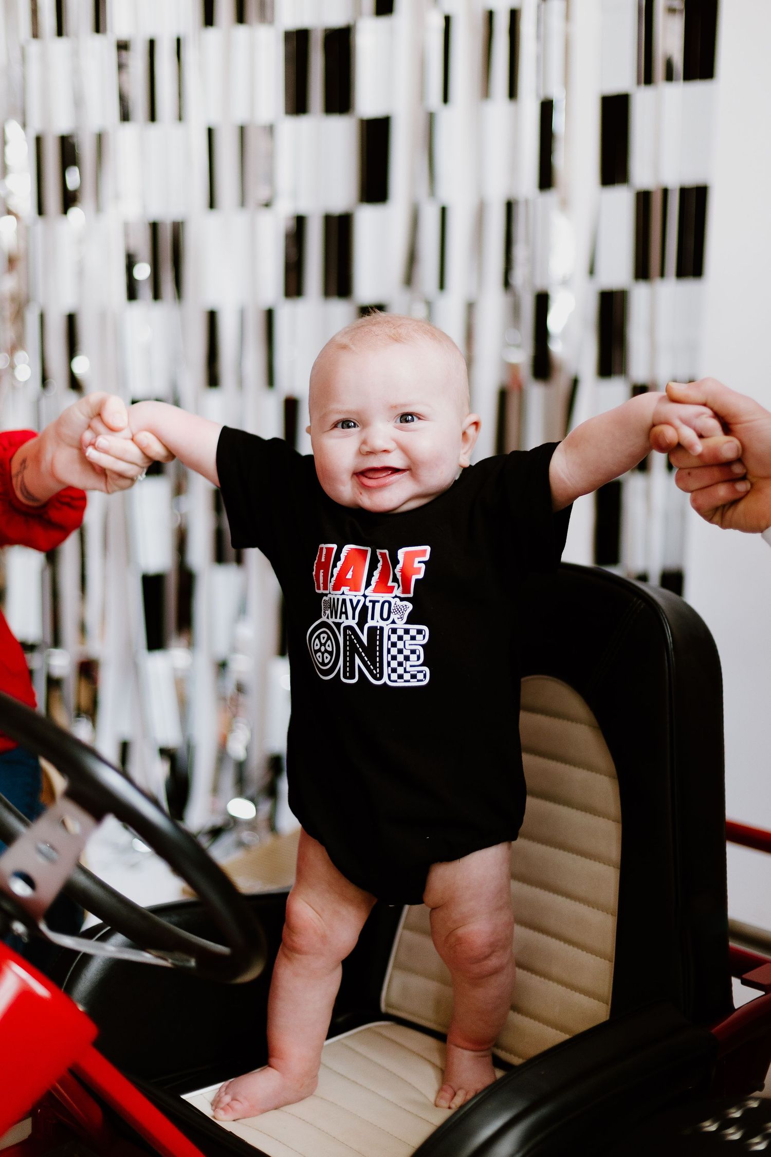 Smiling baby in black race car onesie standing in go-kart at half birthday party.