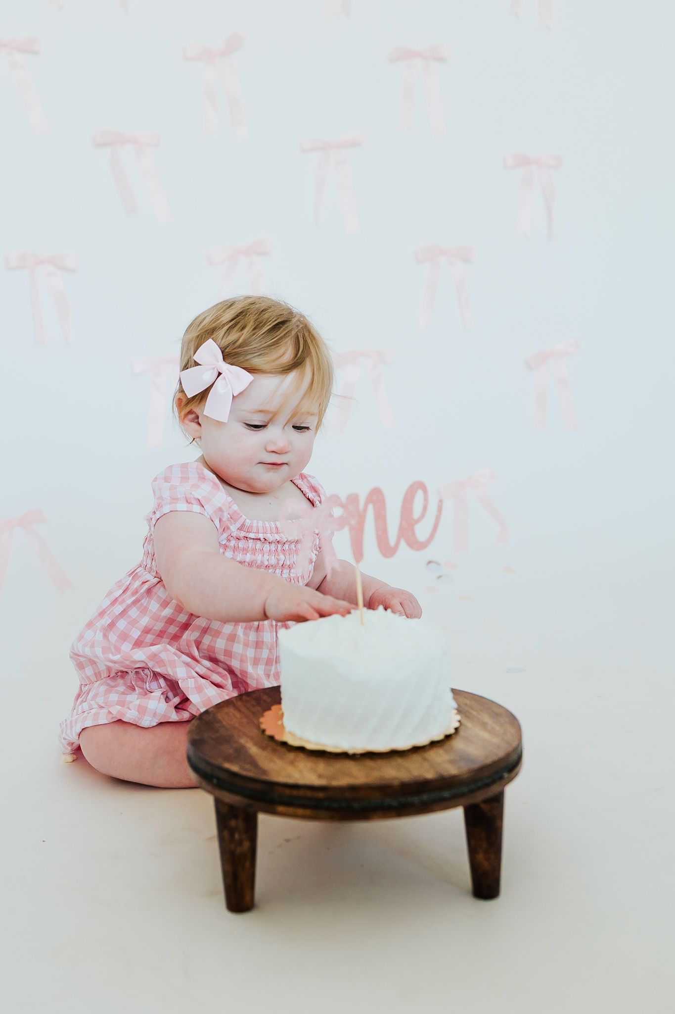 Precious baby girl in pink gingham dress examines first birthday smash cake on small wooden stool.
