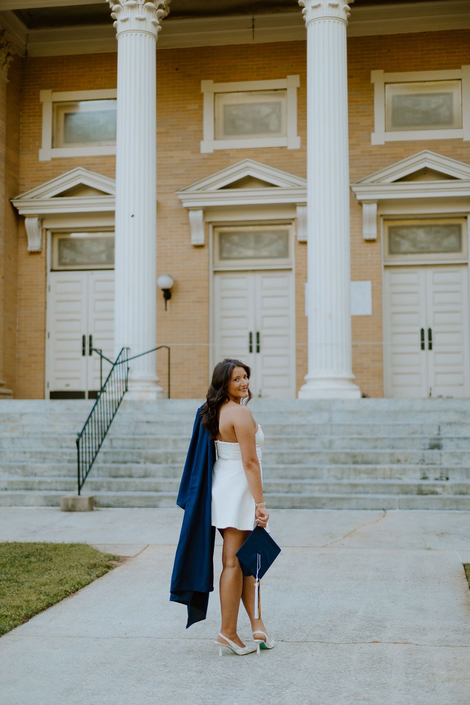 Smiling graduate in white dress holding navy cap and gown outside classical brick building.
