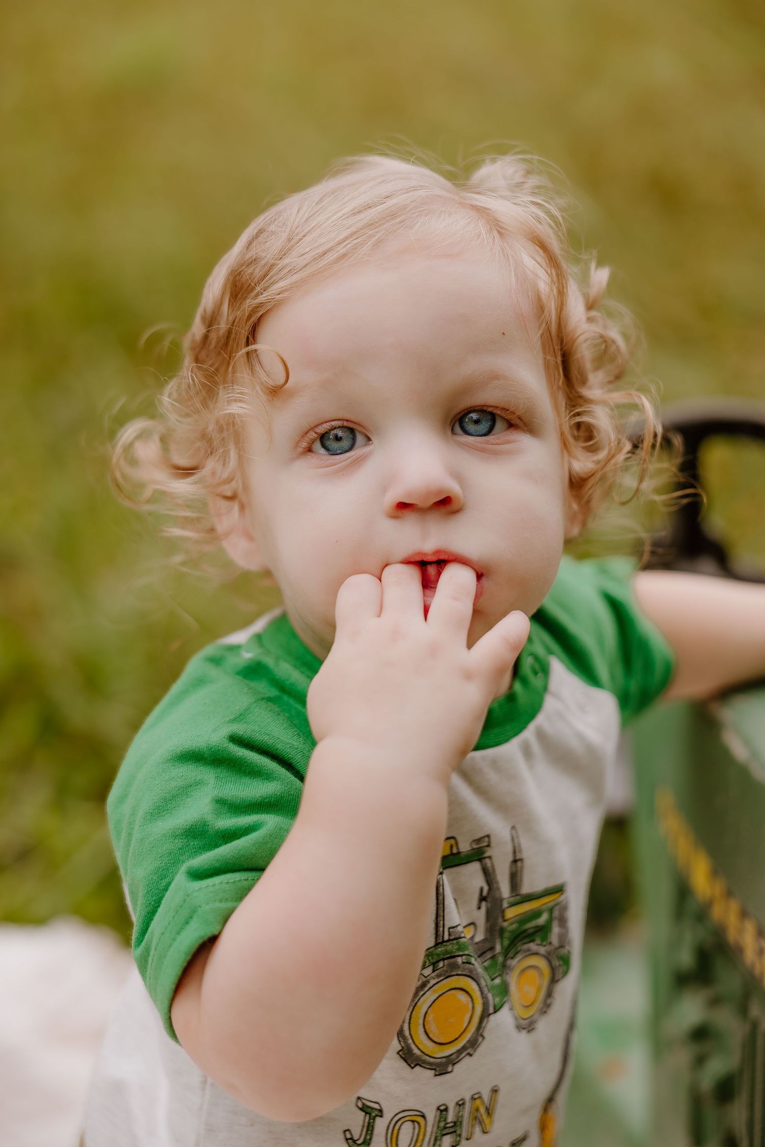 Curly-haired toddler with blue eyes wearing a John Deere tractor shirt, finger in mouth, outdoors on green grass.