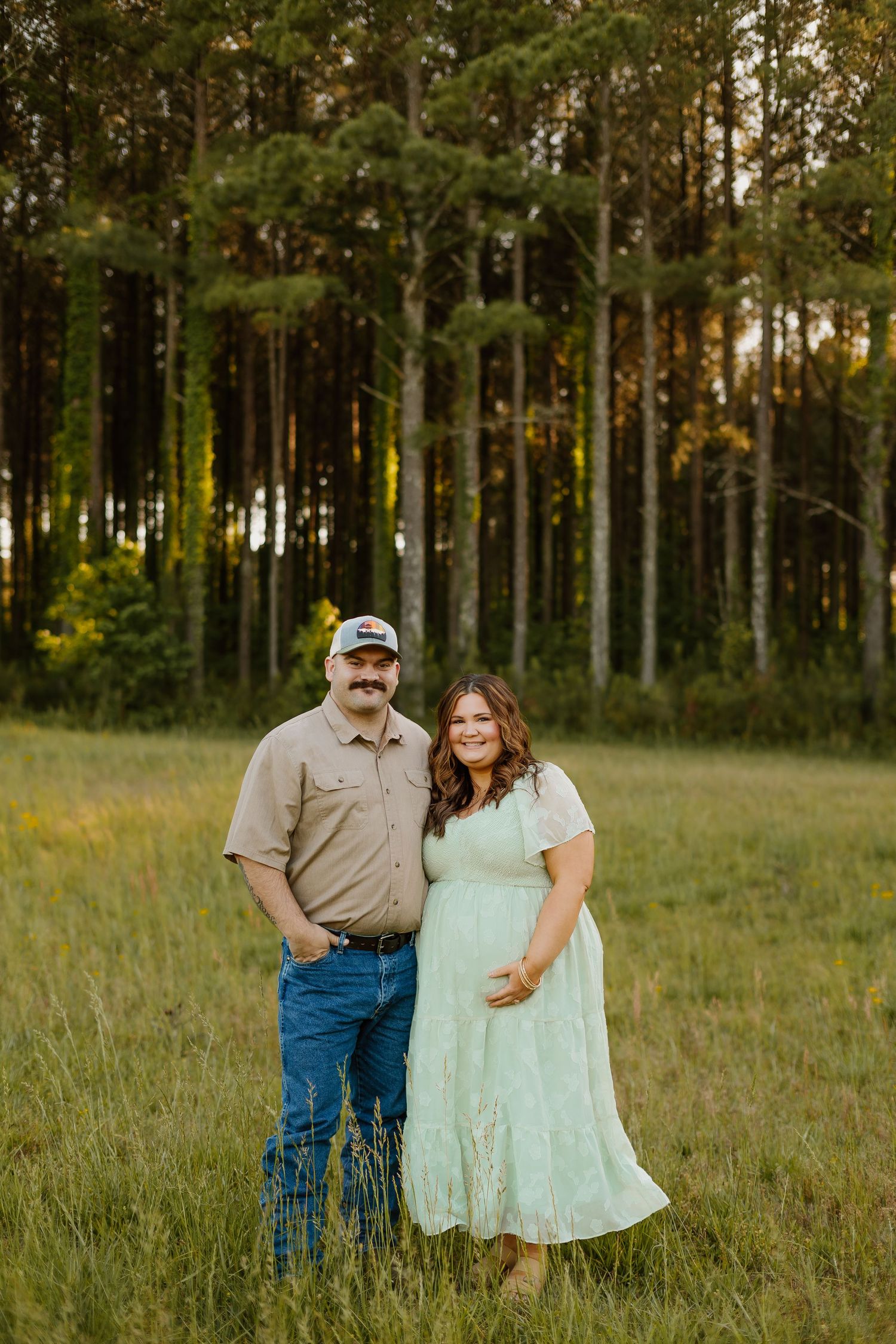 Couple posing in a green meadow with tall pine trees, man in tan shirt and cap, woman in mint dress.