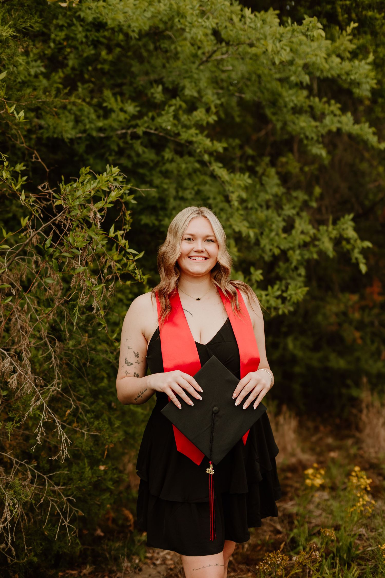 Smiling graduate in black dress with red stole holds cap outdoors among lush green trees.