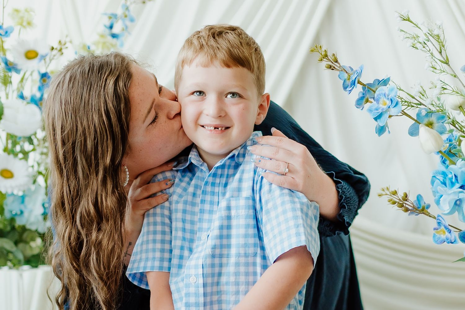 A mother lovingly kisses her smiling young boy in a blue shirt against a floral white backdrop.