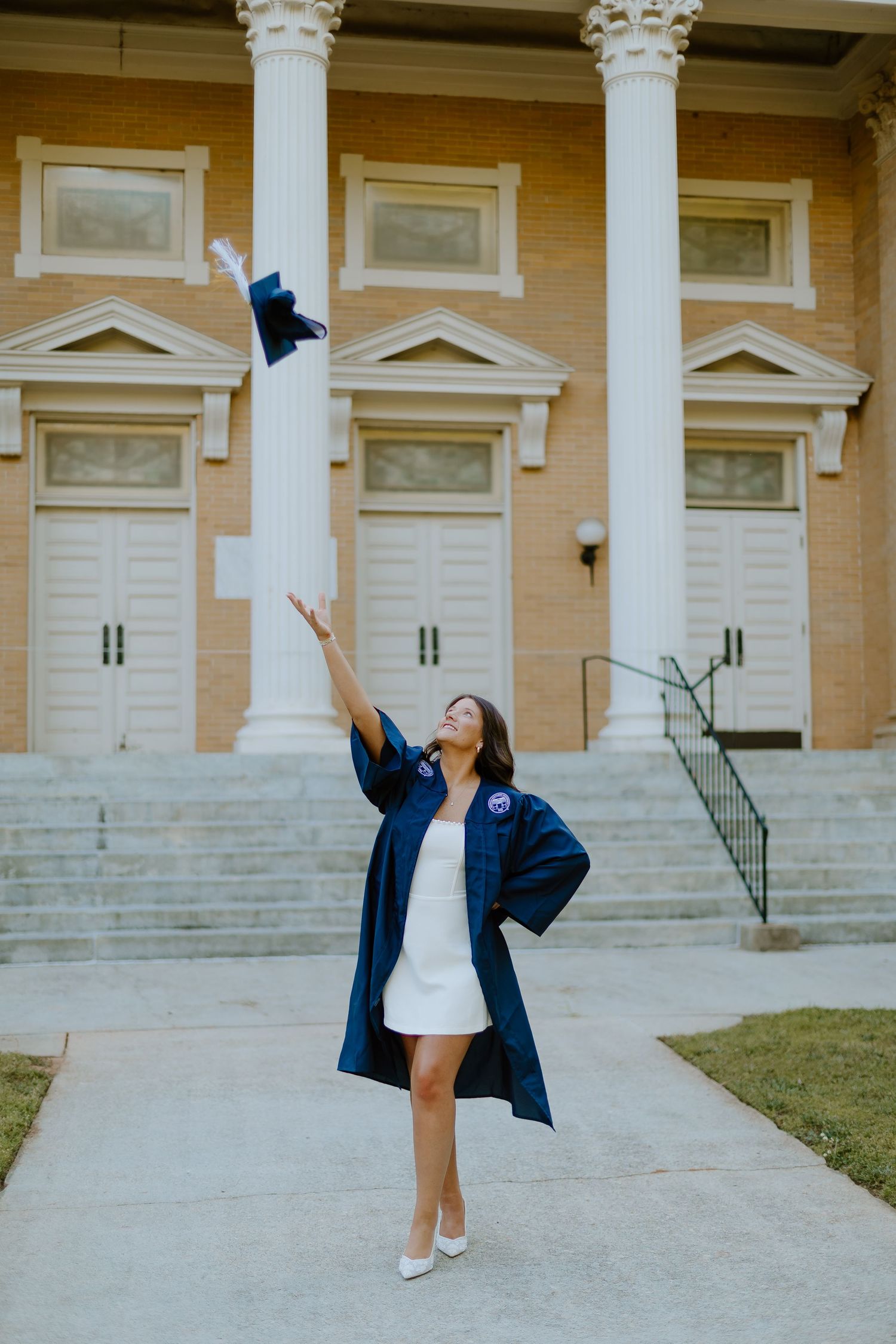 Graduate in navy gown tossing mortarboard cap in air in front of university building.