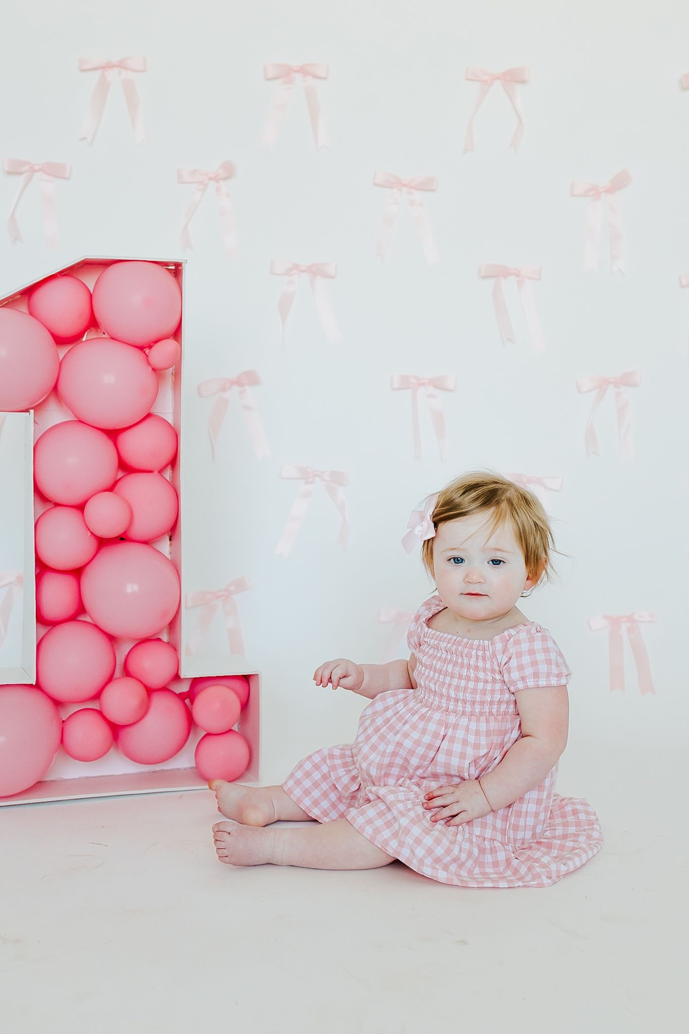 Cute baby girl in pink gingham dress sits beside large number one balloon display for first birthday.