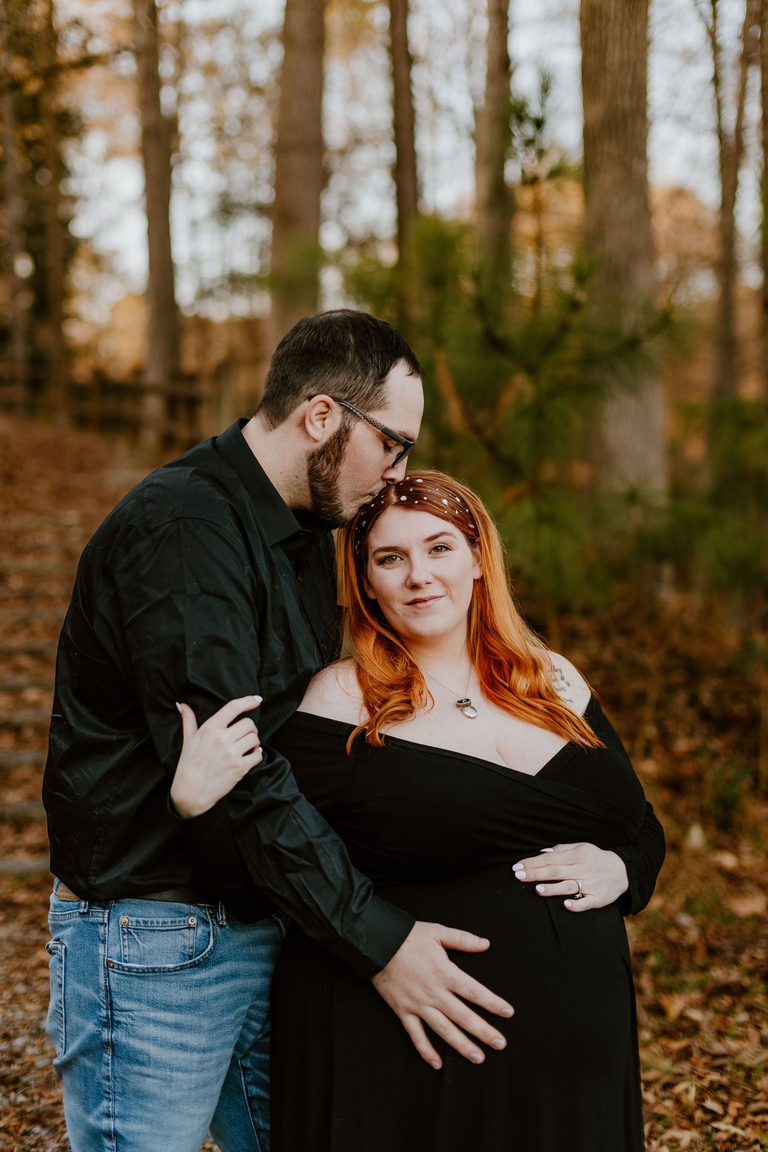 Couple maternity photo in autumn forest, man kissing pregnant woman with red hair wearing black off-shoulder dress.