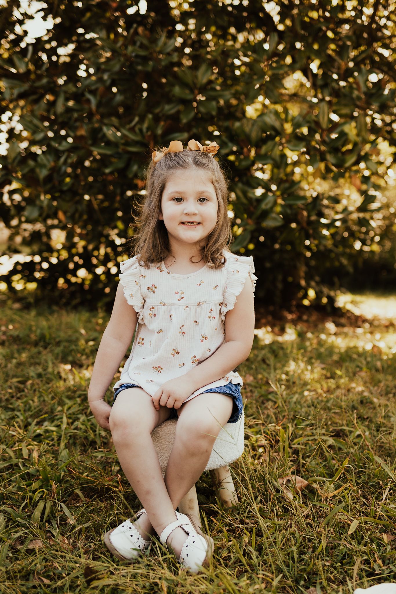 Adorable young girl in a floral dress sitting on a small stool outdoors in a sunny garden.