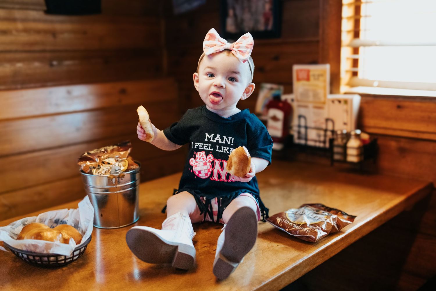 Baby girl in first birthday outfit sits on restaurant table surrounded by food baskets with bow headband.