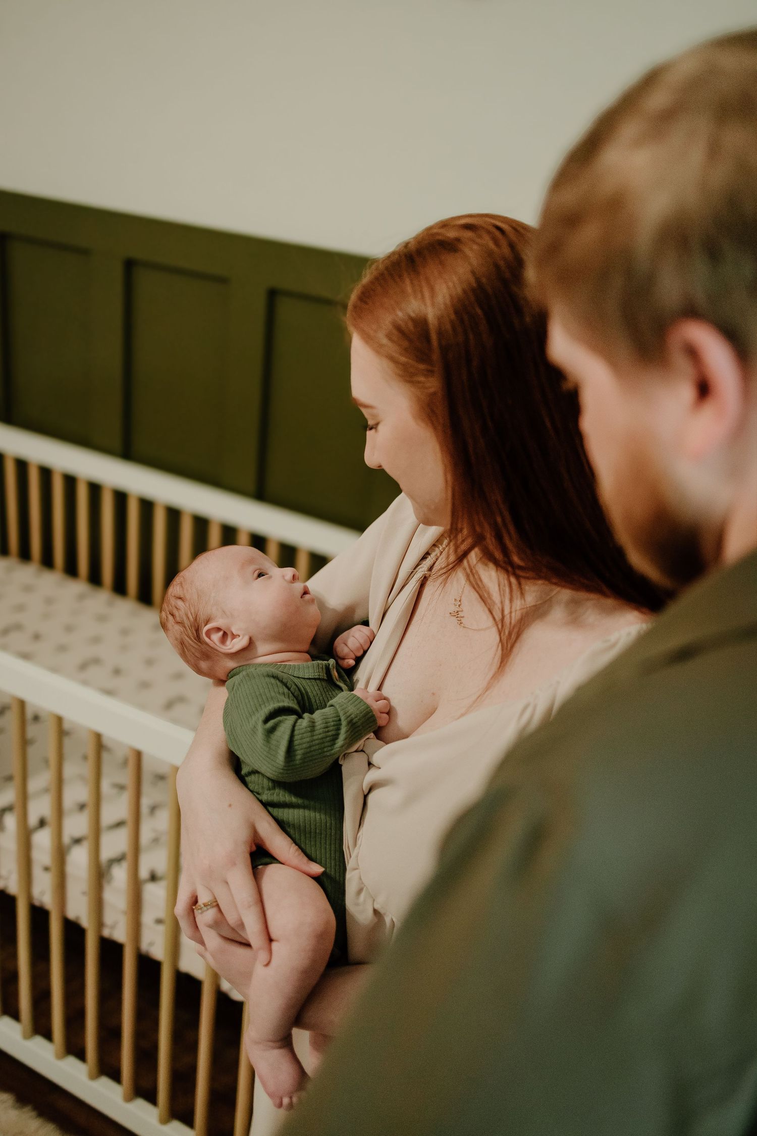 Loving mother holding newborn baby in green onesie with father beside her near crib.