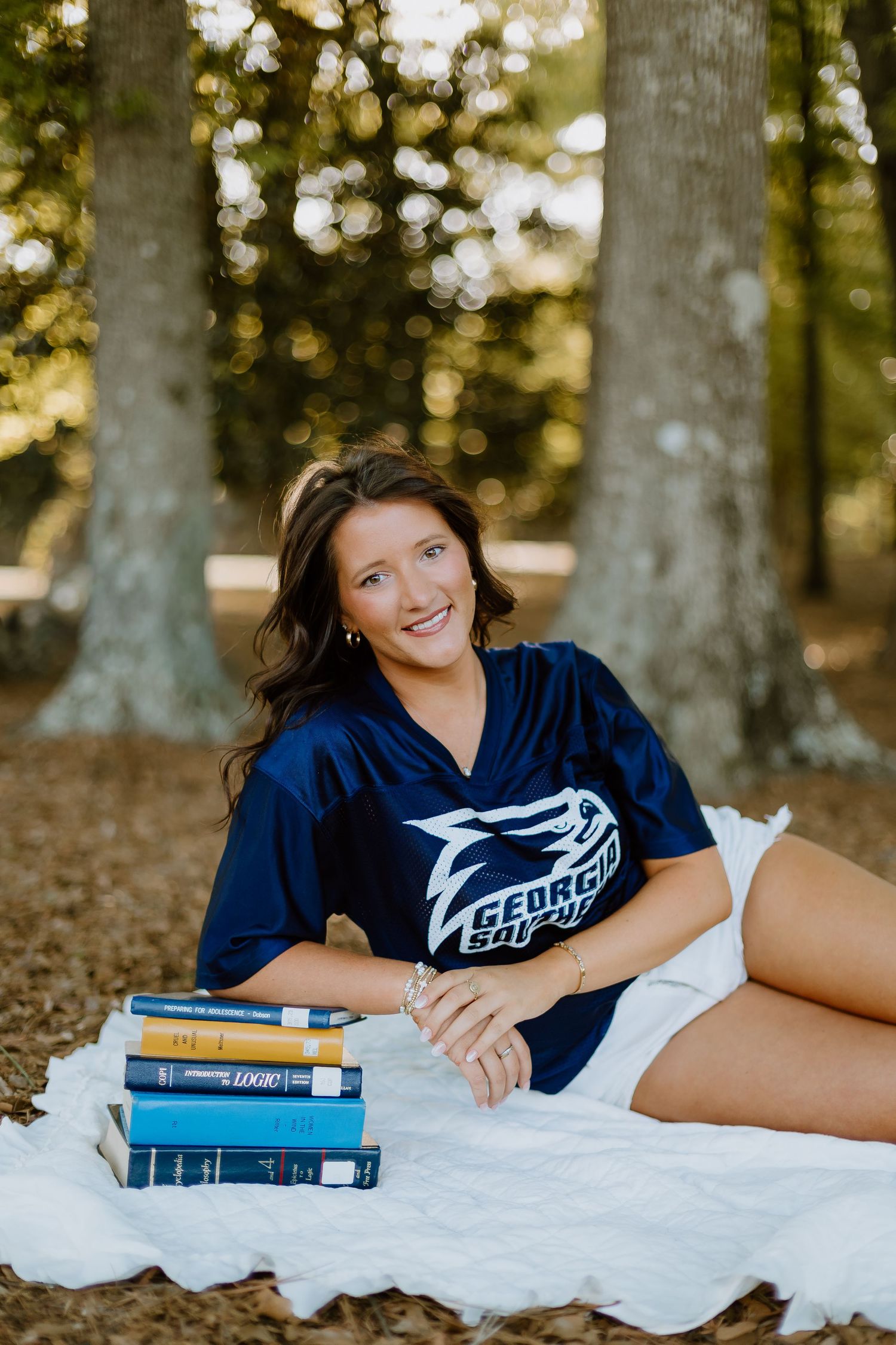 Smiling woman in Georgia Southern jersey lying on white blanket with books among trees.