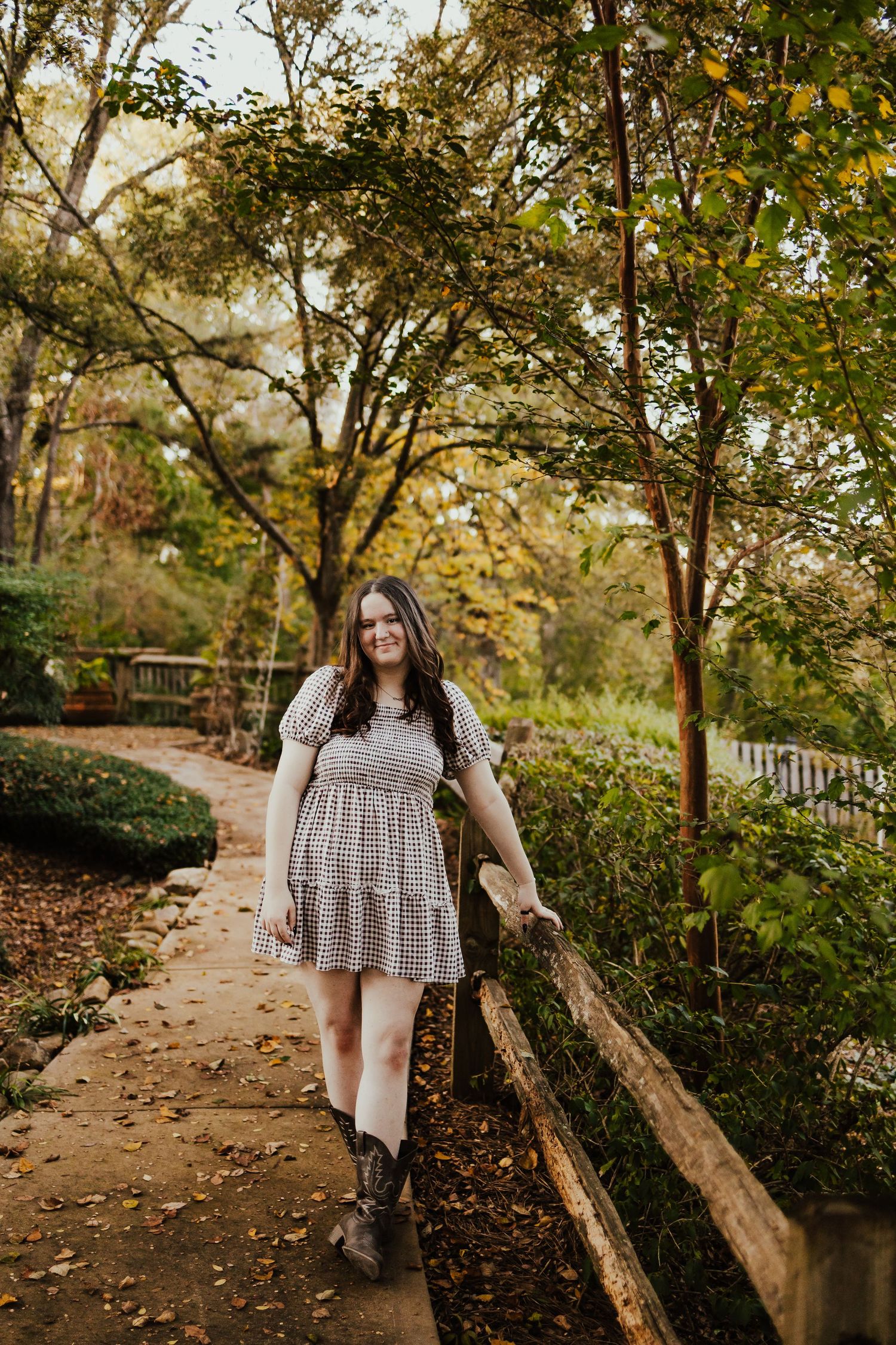 Young woman in gingham dress smiling while walking along garden path with autumn foliage backdrop.