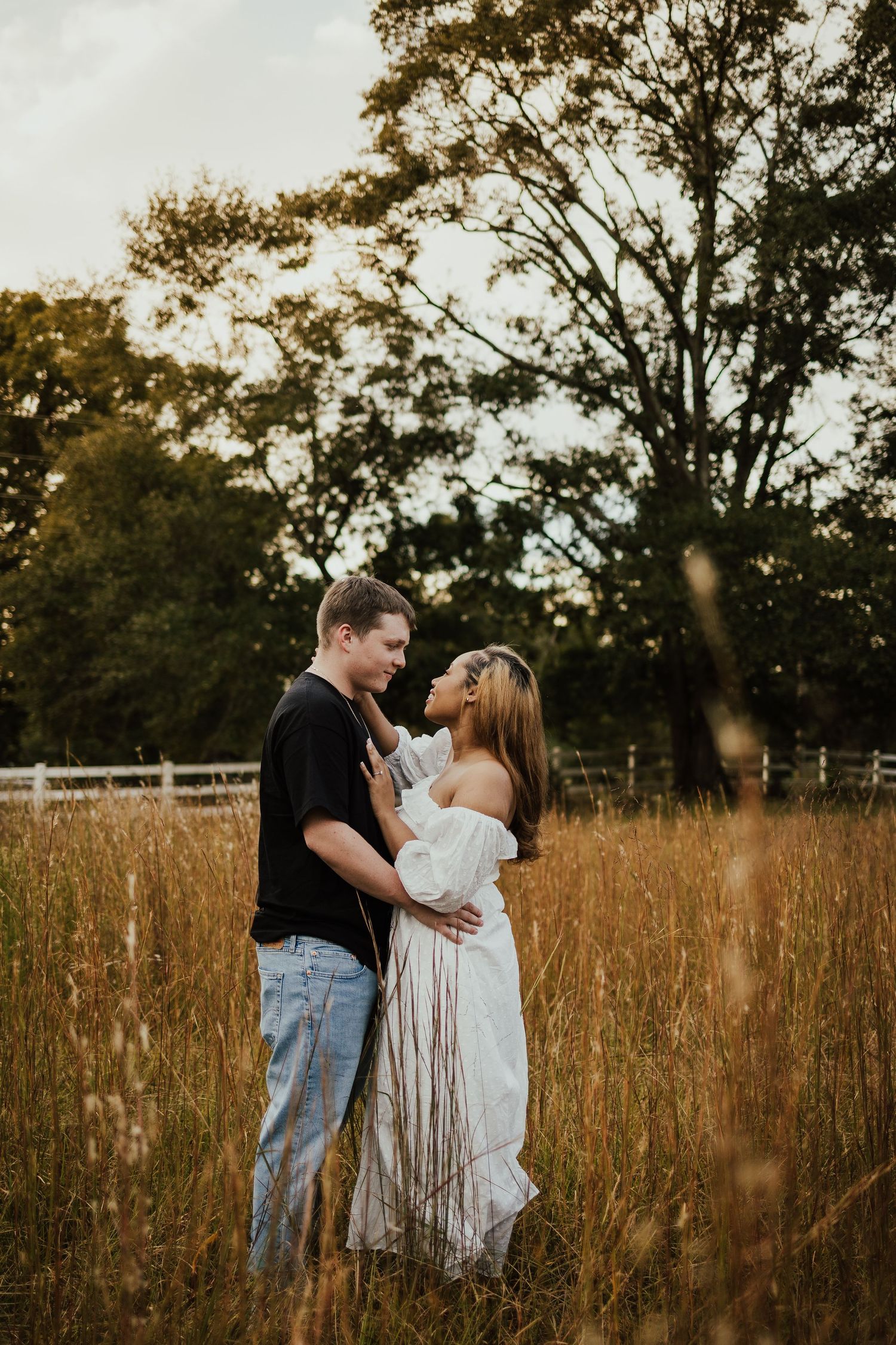 Young couple holding each other in a tall golden grass field near a white fence at dusk.