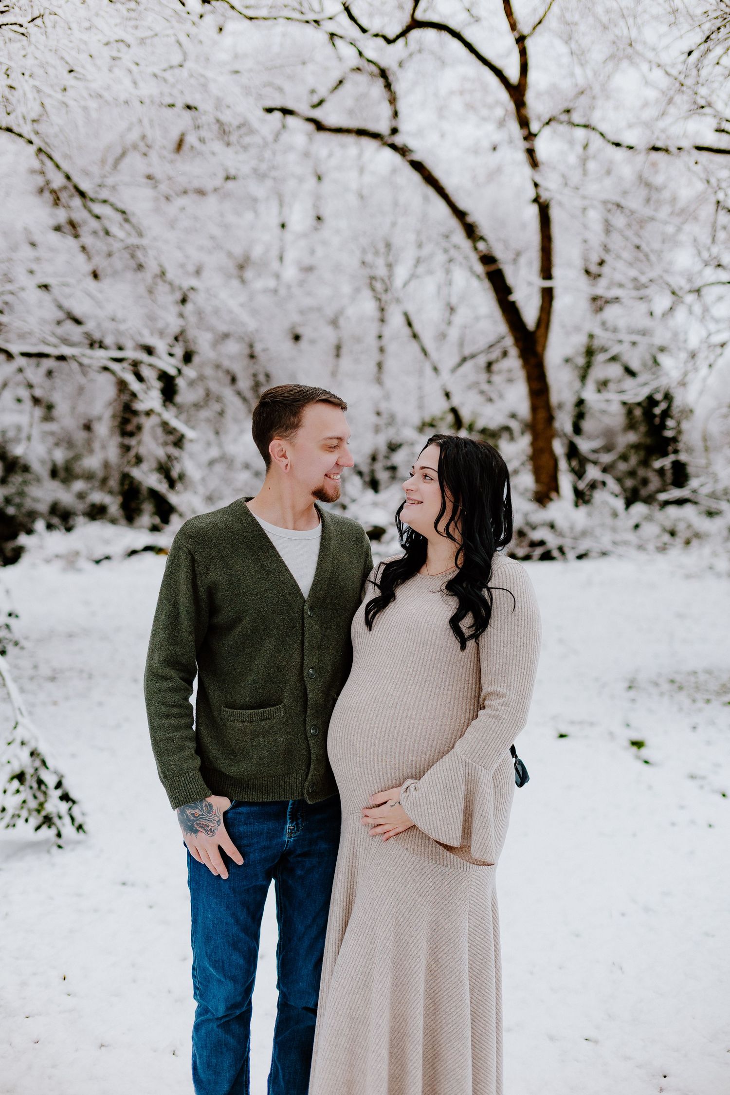 Expectant couple shares tender moment in snow-covered forest, woman in knit maternity dress.