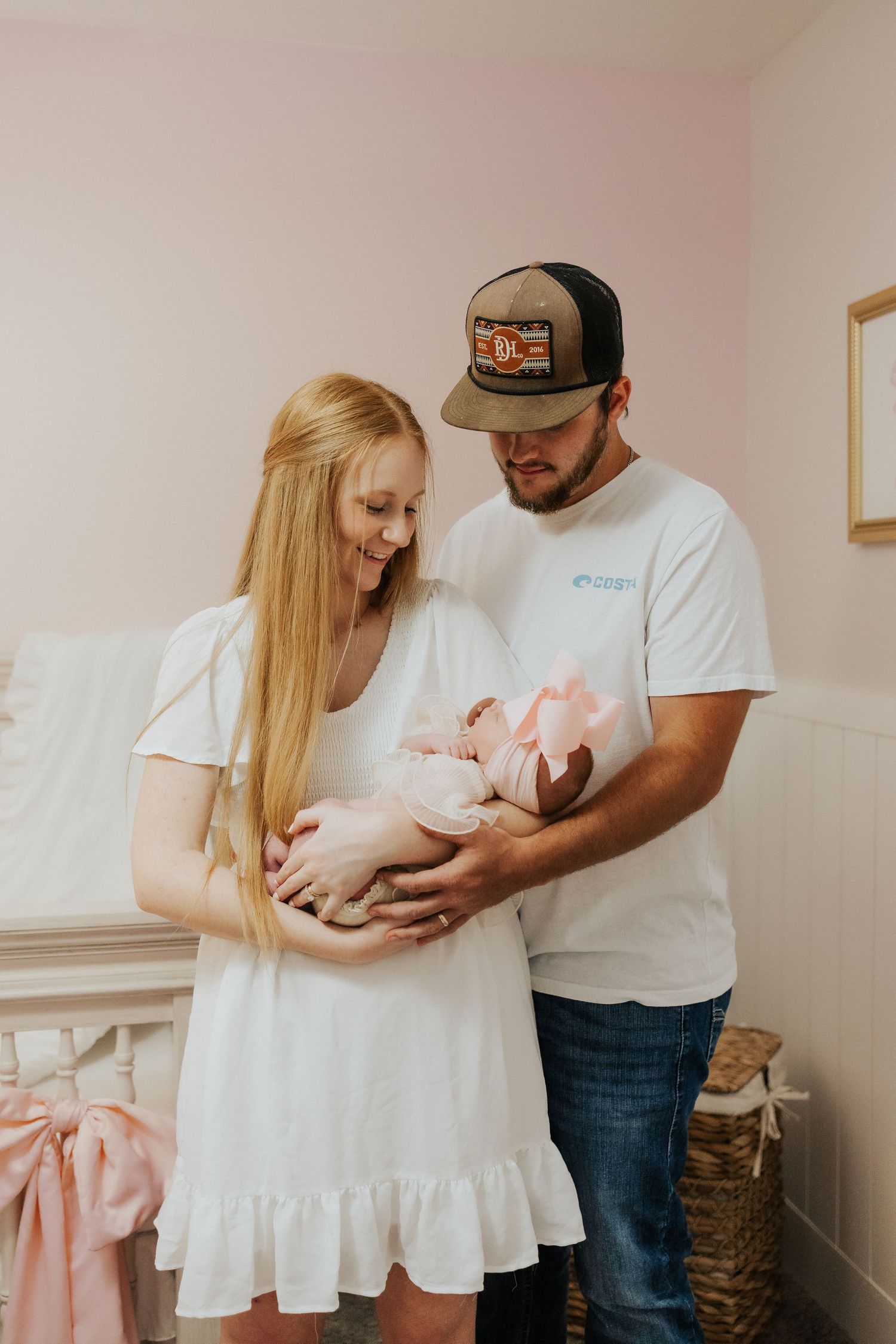 Happy new parents in white outfits tenderly hold their newborn baby dressed in pink in a soft nursery.