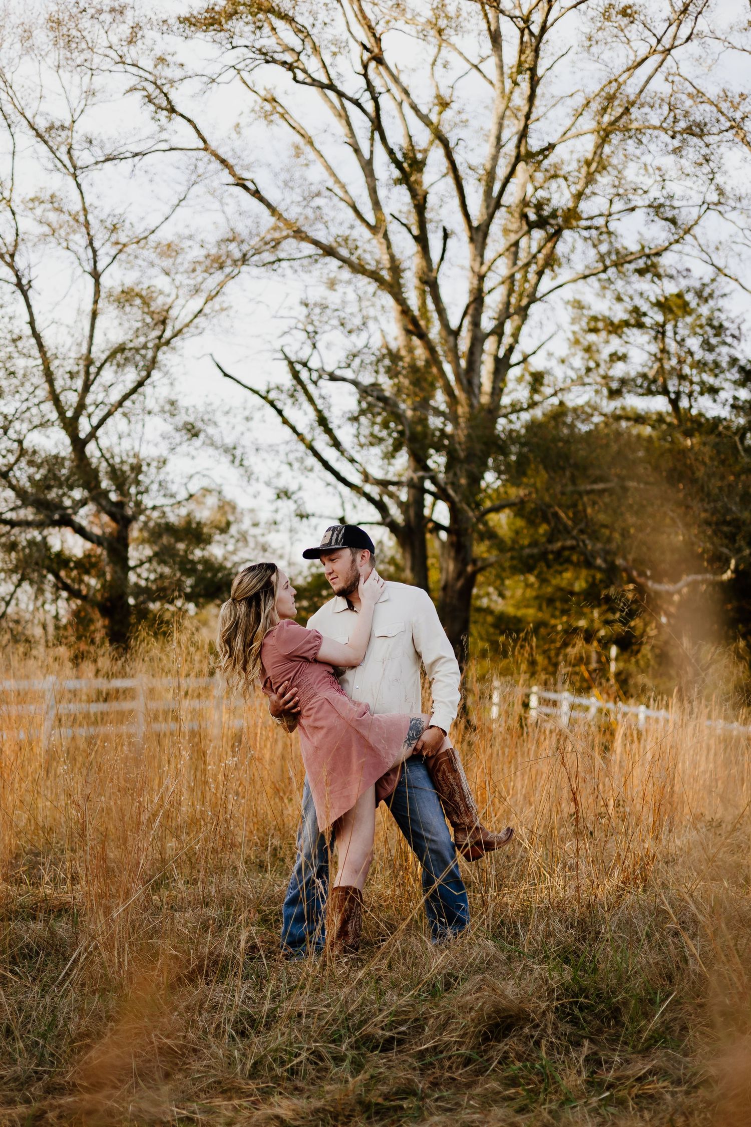 Couple embracing romantically in a golden field of tall grass near a white fence at sunset.
