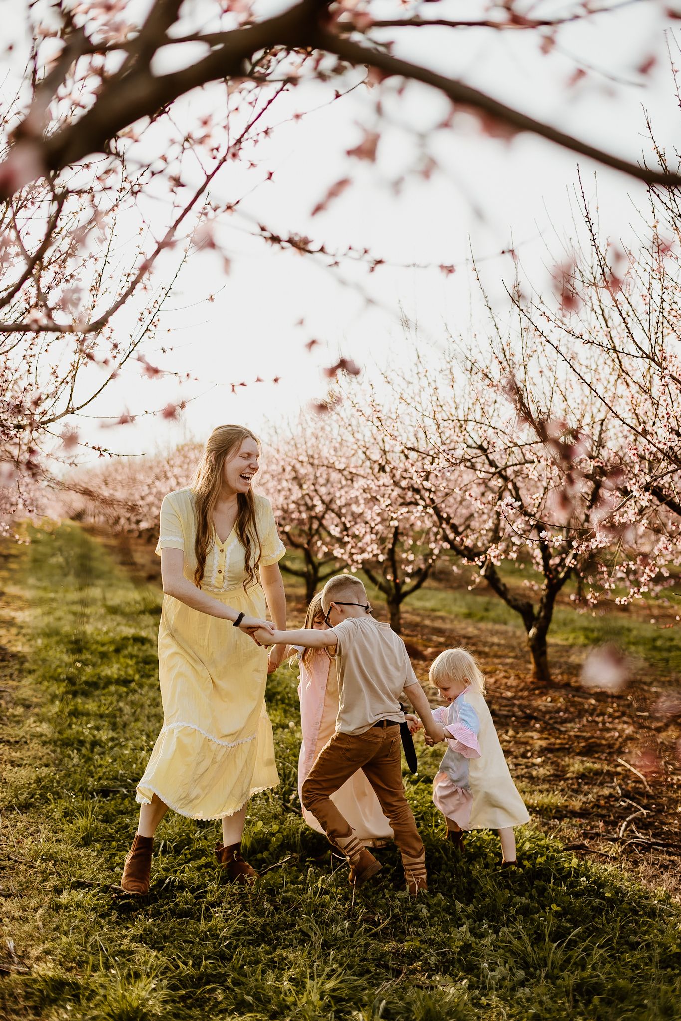 Woman in yellow dress laughing with two children playing in a blooming pink cherry orchard during spring.