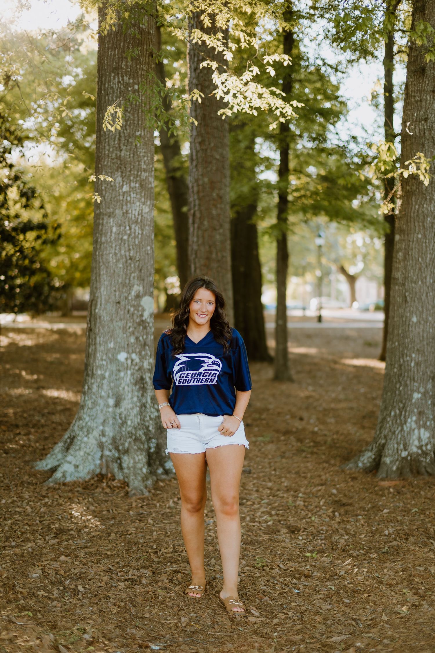 Young woman in Georgia Southern jersey and white shorts posing among tall trees outdoors.