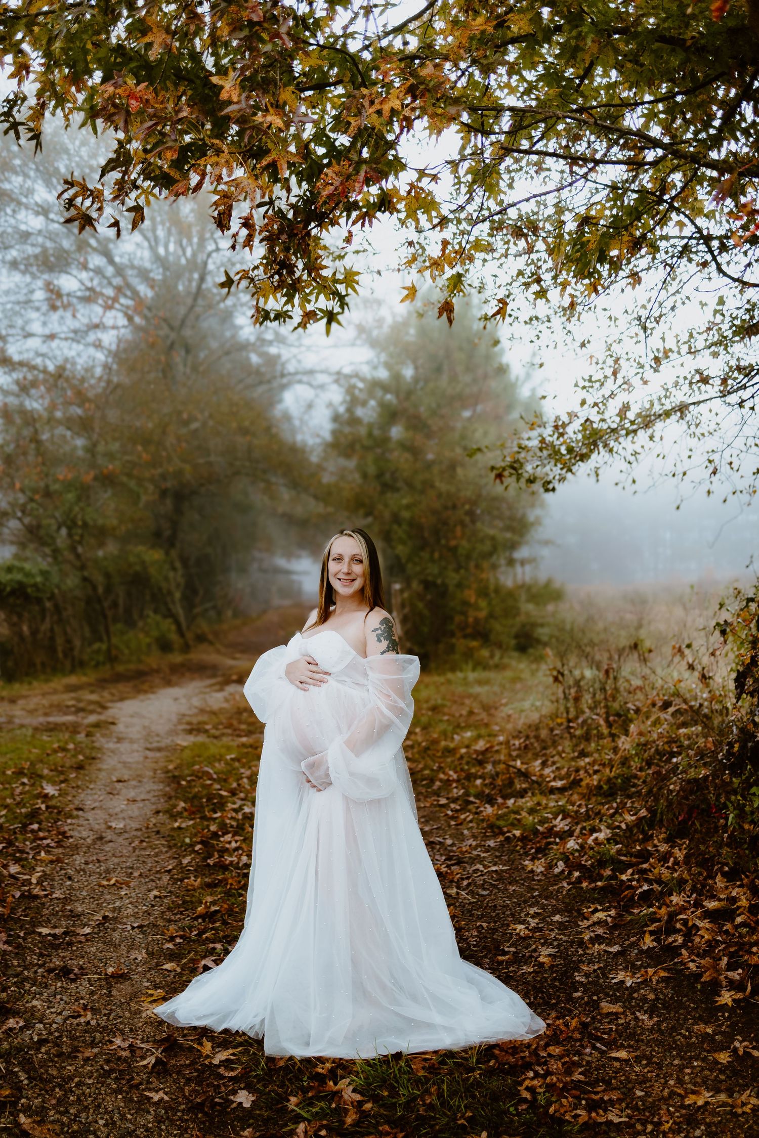 Pregnant woman in elegant white gown posing on foggy autumn forest path.