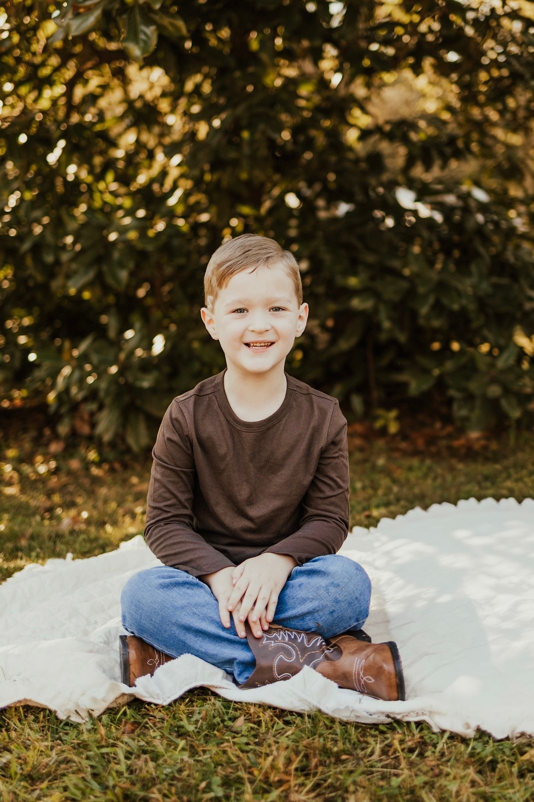 Smiling young boy in a brown shirt and jeans sitting cross-legged on a white blanket outdoors.
