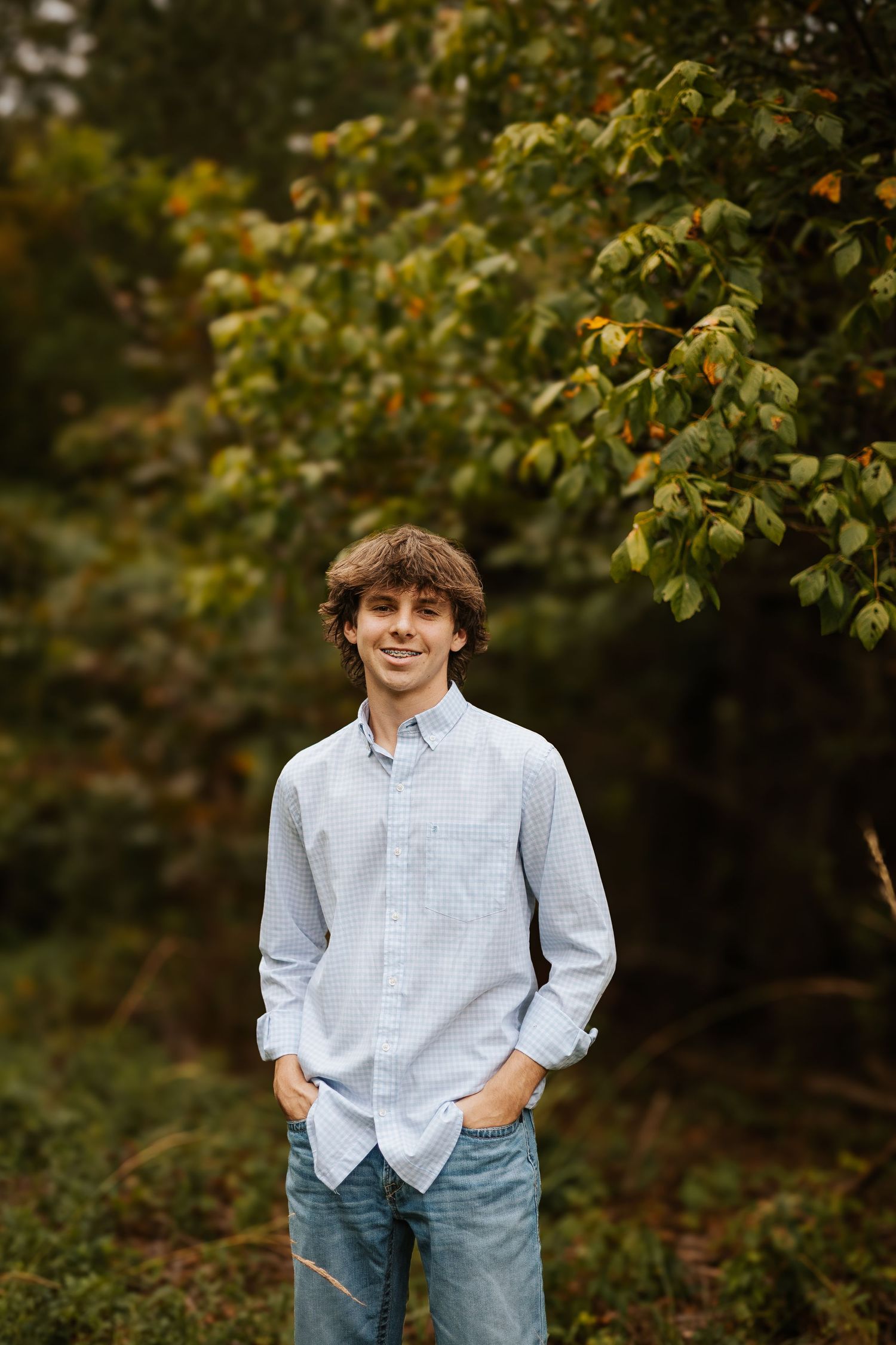 Smiling senior boy in light blue button-down shirt standing before colorful autumn foliage outdoors.