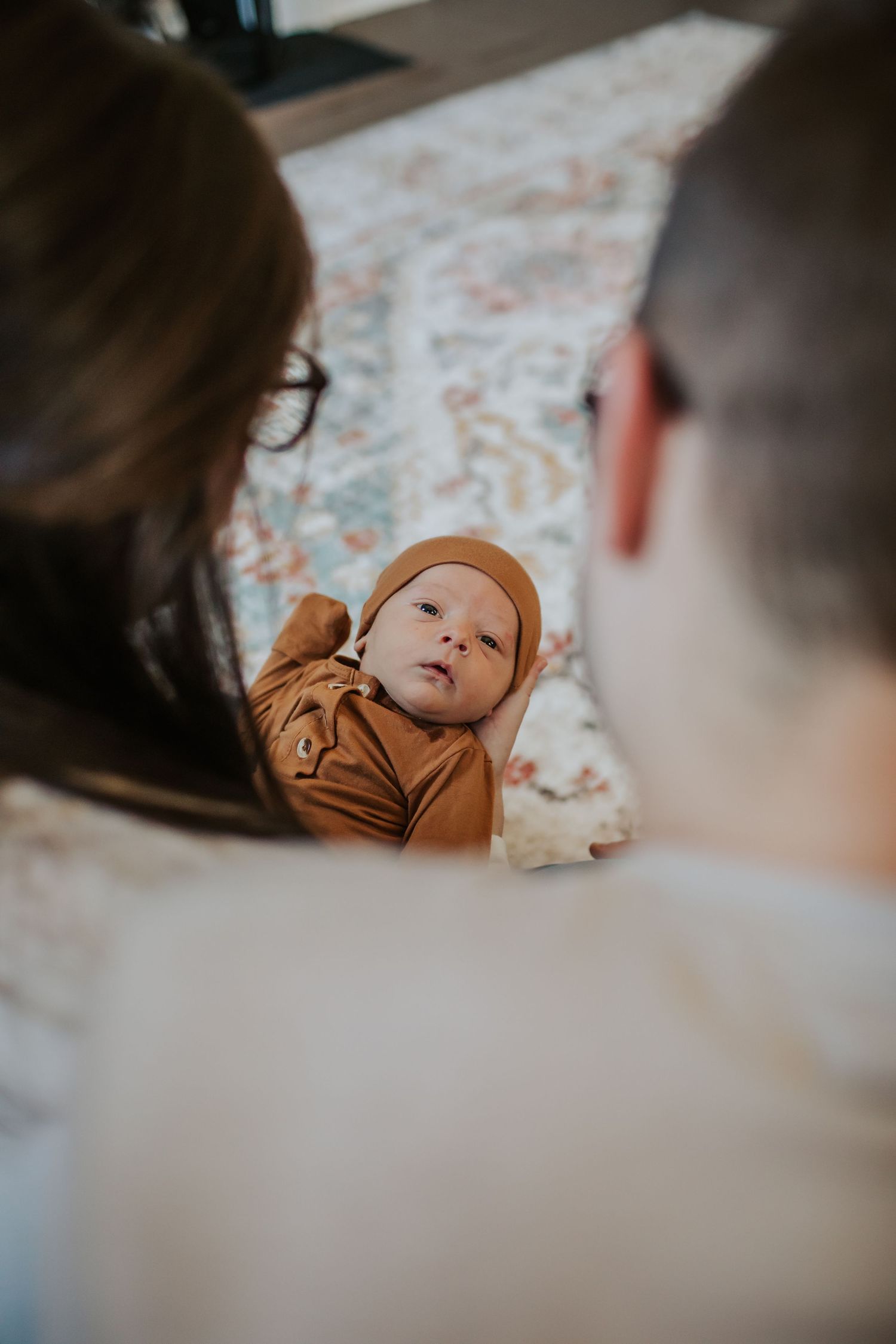 Newborn baby in brown outfit looks up at parents from a floral rug during tummy time.