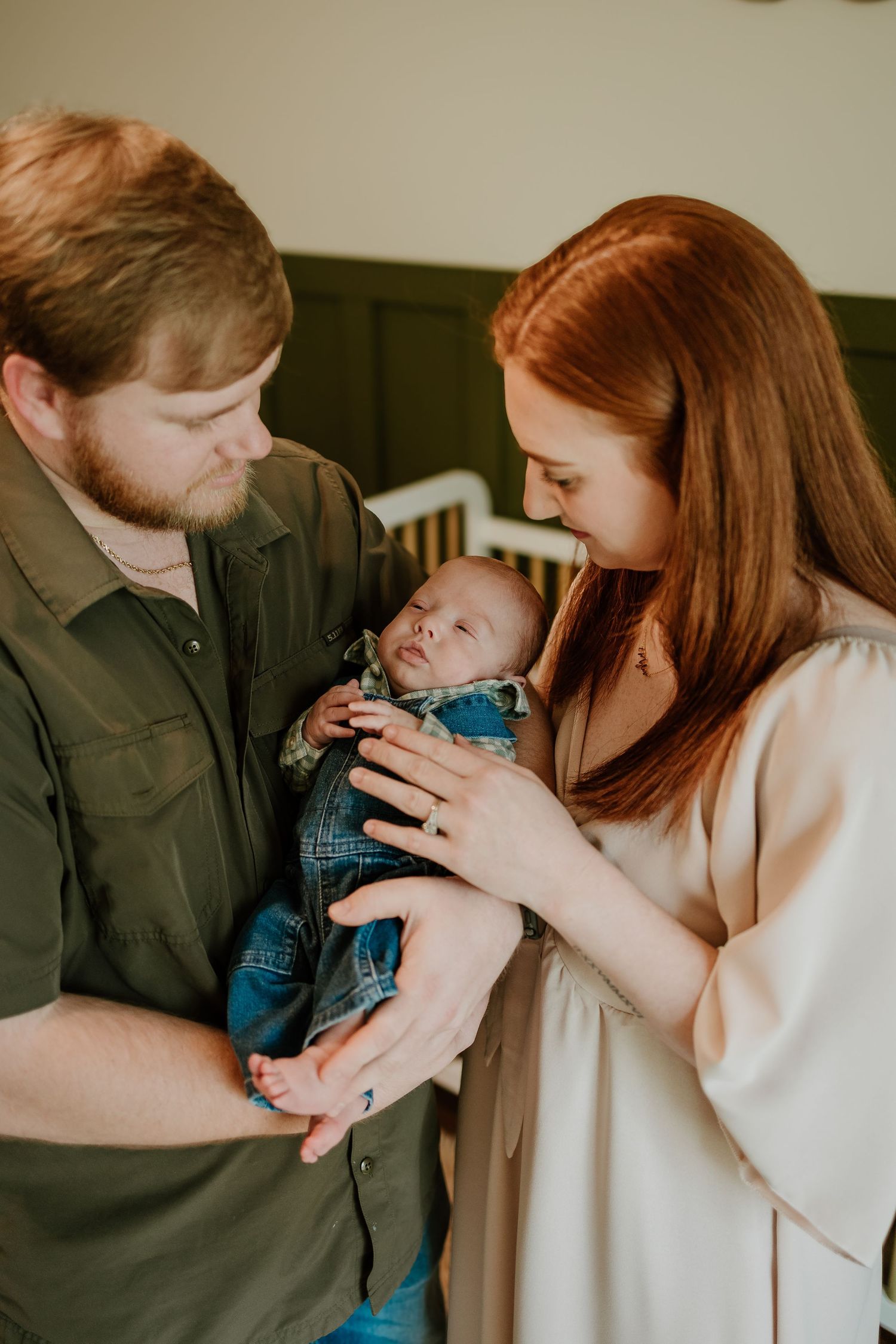 New parents lovingly gazing at their sleeping newborn baby dressed in denim overalls.