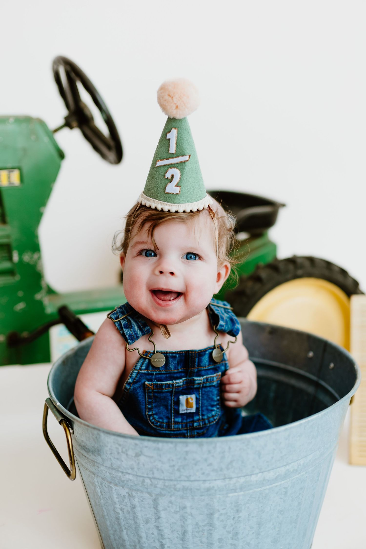 Baby in denim overalls wearing half birthday party hat sitting in galvanized bucket with tractor.