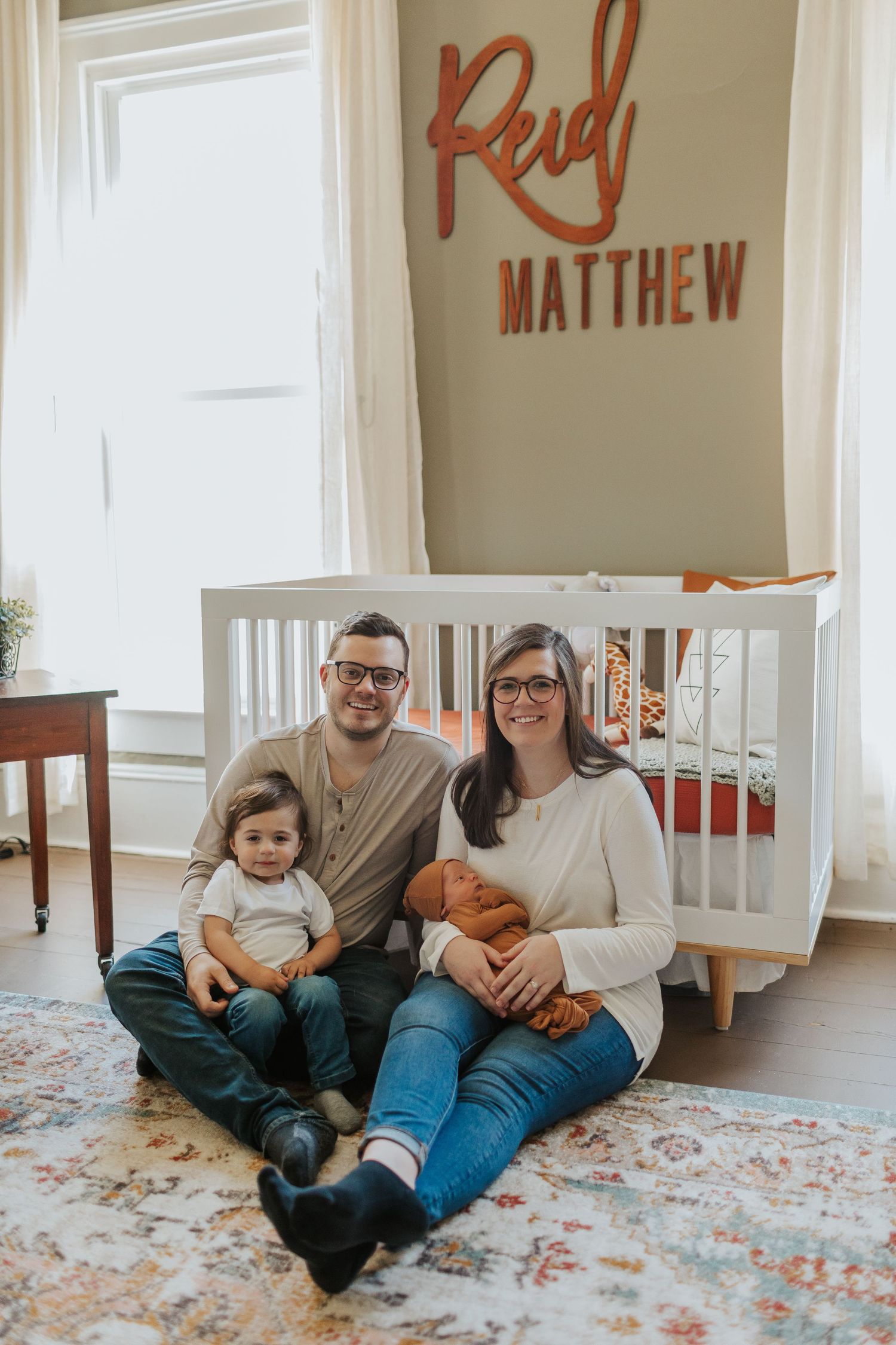 Family of three sitting in nursery beside white crib with Reid Matthew name sign on wall.
