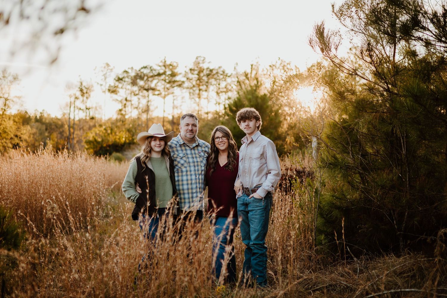 Family of four posing together in a golden field of tall grass at sunset with pine trees behind them.