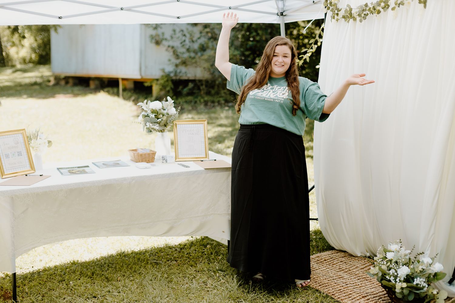 Woman in teal sweater and black skirt posing with open arms at an outdoor wedding vendor booth.