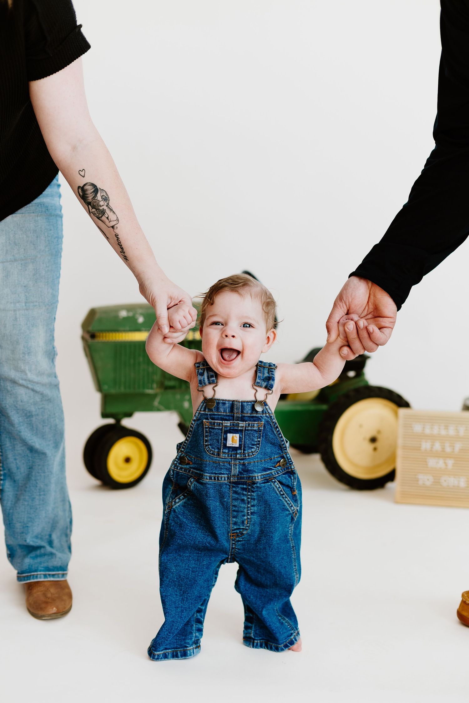 Smiling baby in Carhartt overalls standing with parent support near green John Deere tractor.
