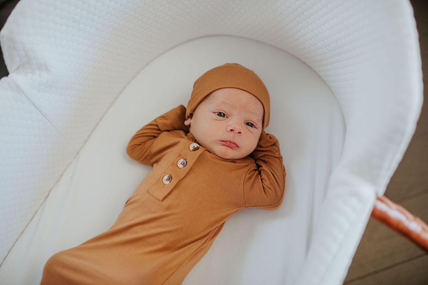 Newborn baby in caramel knit gown and hat lying in white bassinet during newborn photo session.