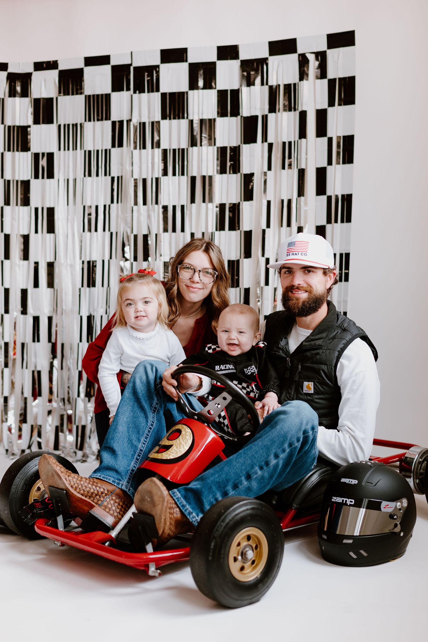 Family of four poses together in red go-kart against checkered black and silver fringe backdrop.