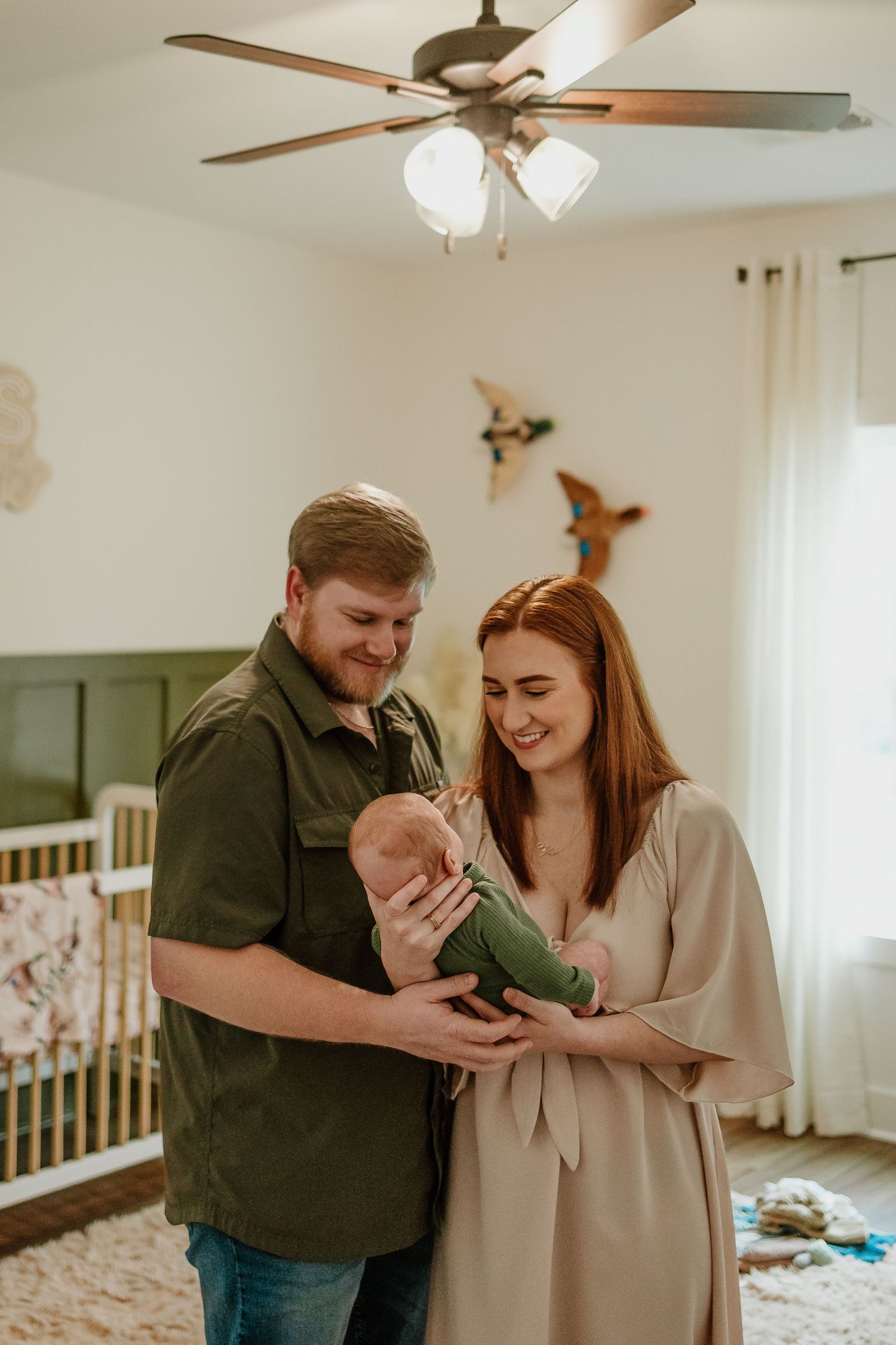 Smiling new parents holding their newborn baby in a green onesie in a decorated nursery.