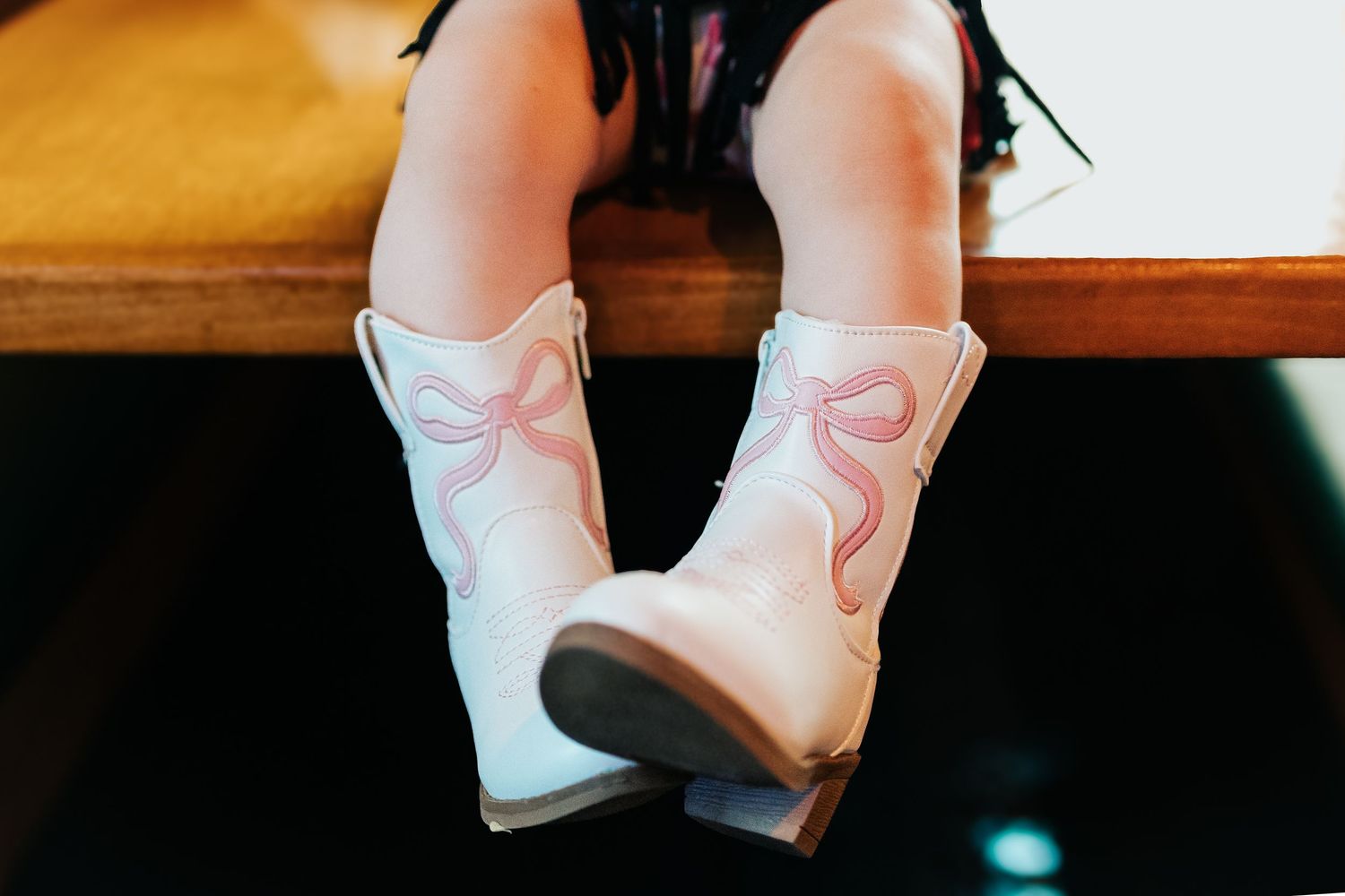 Close-up of toddler girl wearing white cowboy boots with pink bow detail sitting on wooden bench.