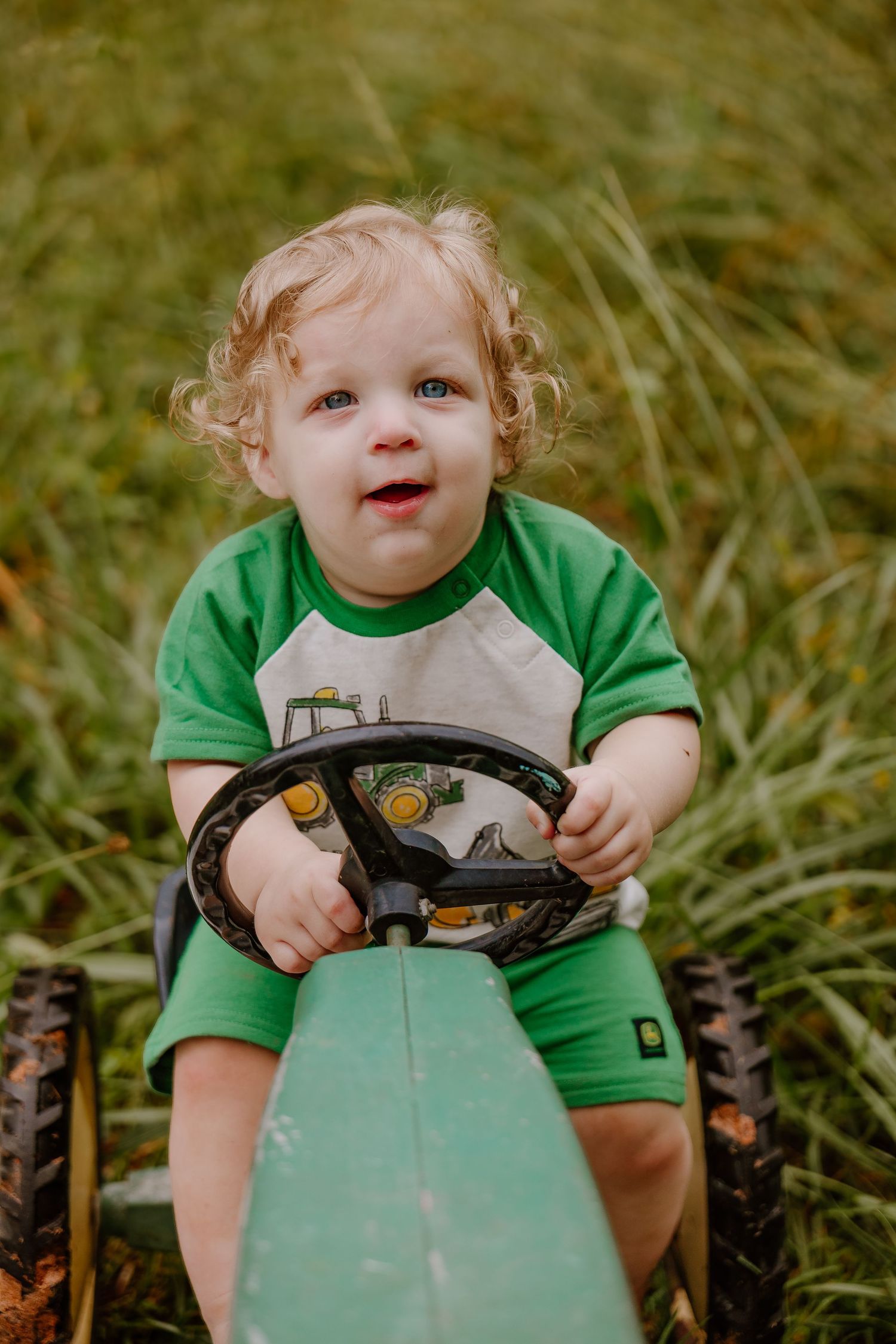 Smiling toddler in green John Deere outfit sitting on toy tractor in grassy field.