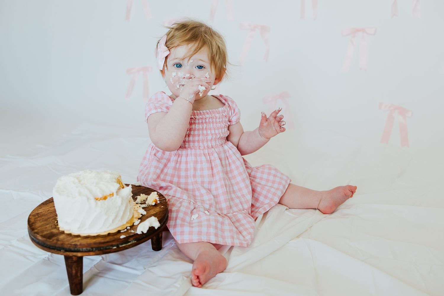 Sweet baby girl in pink gingham dress sits beside smash cake on rustic wooden stool.