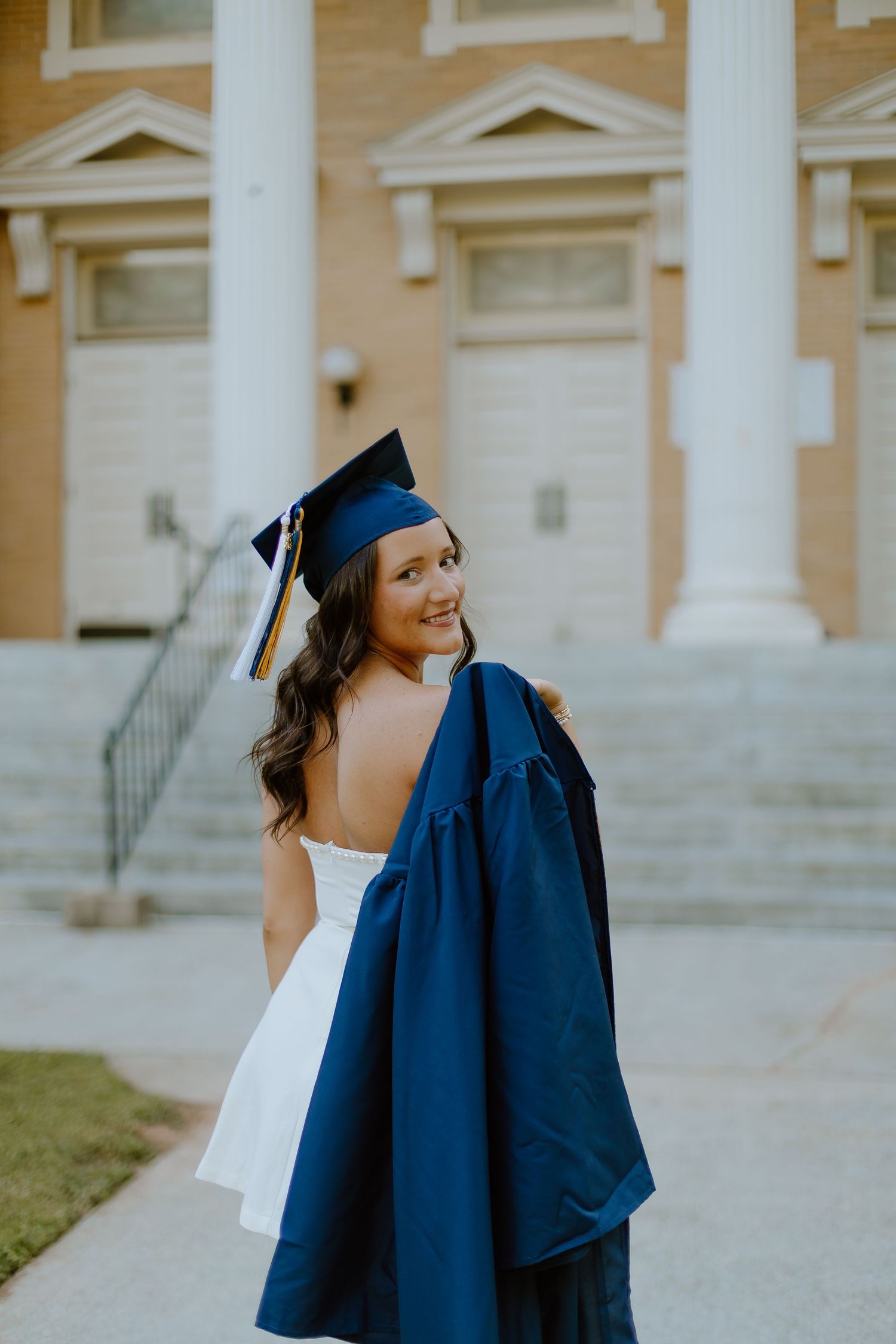 Graduate in navy cap and gown looking over shoulder smiling outside brick college building.
