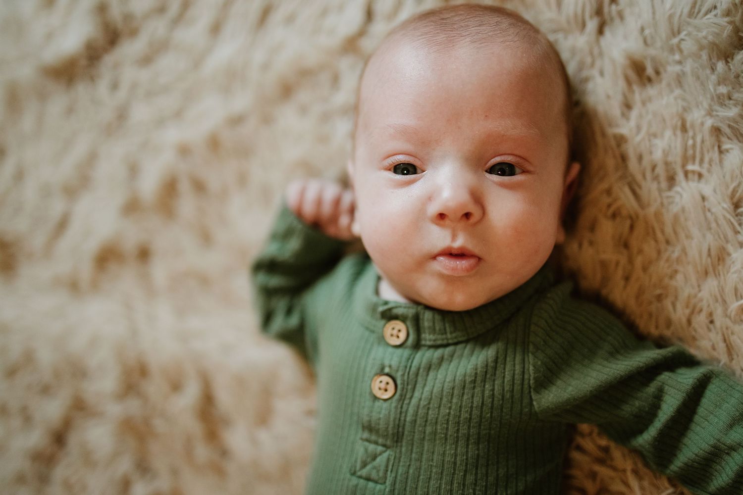 Adorable newborn baby boy in a green ribbed onesie lying on a soft fluffy cream rug.