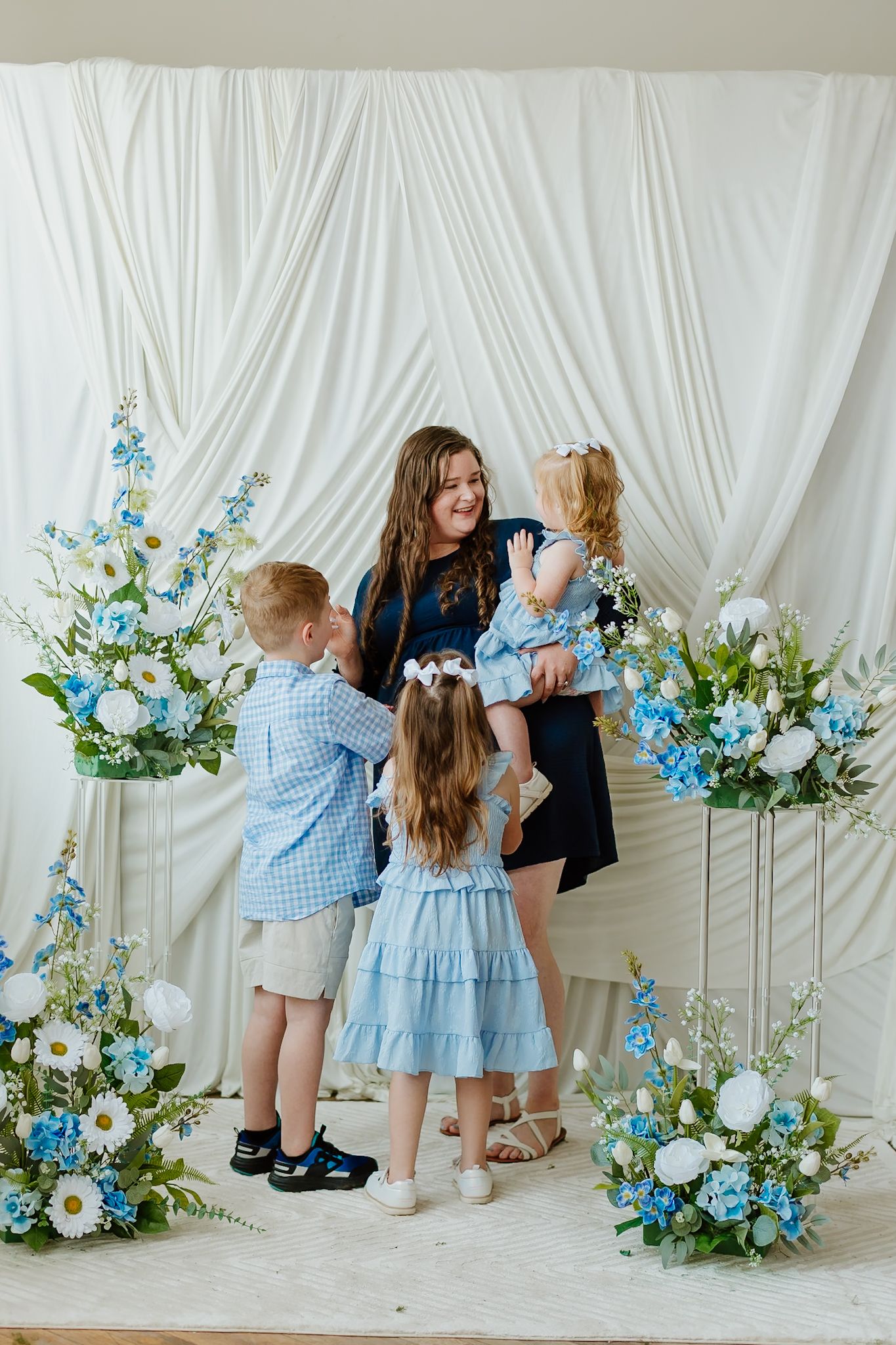 A mother in navy dress holds toddler surrounded by children in blue outfits near white drape with blue floral arrangements.