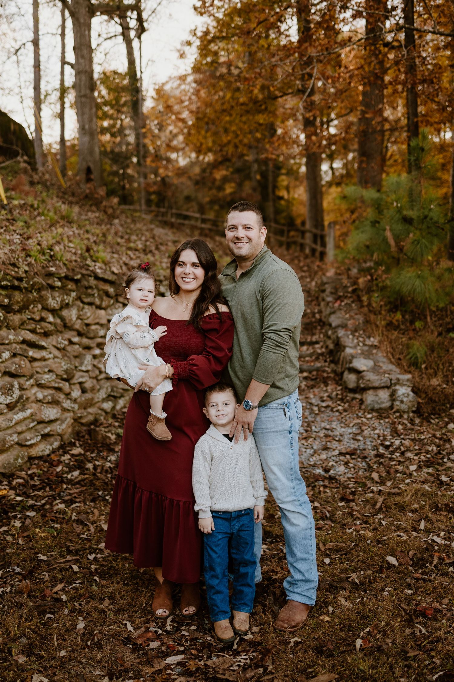 Family of four poses in autumn forest, mother in red dress holding toddler girl beside father and young son.