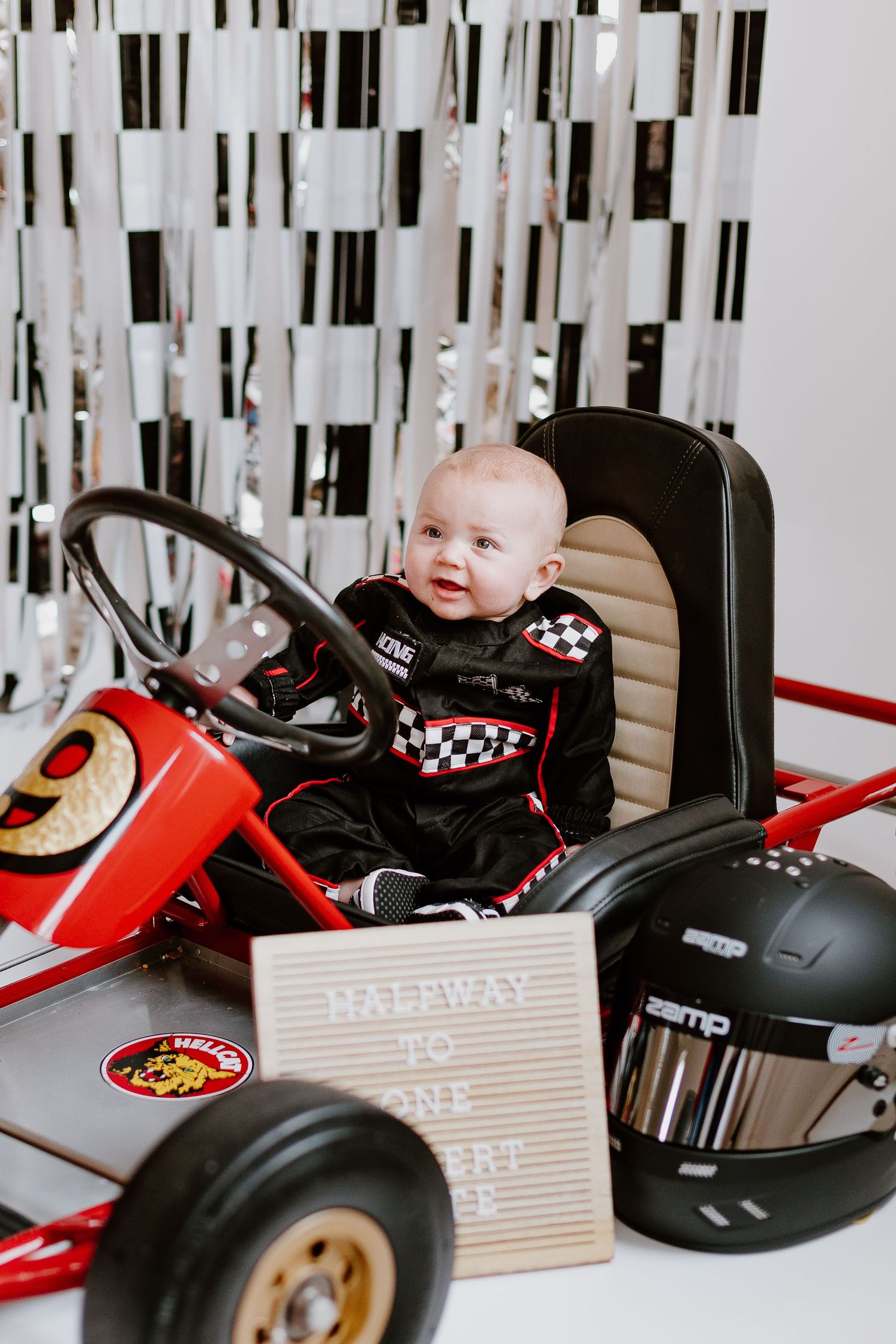 Baby in racing suit sits in red go-kart with helmet and letterboard sign for 6-month milestone photo.