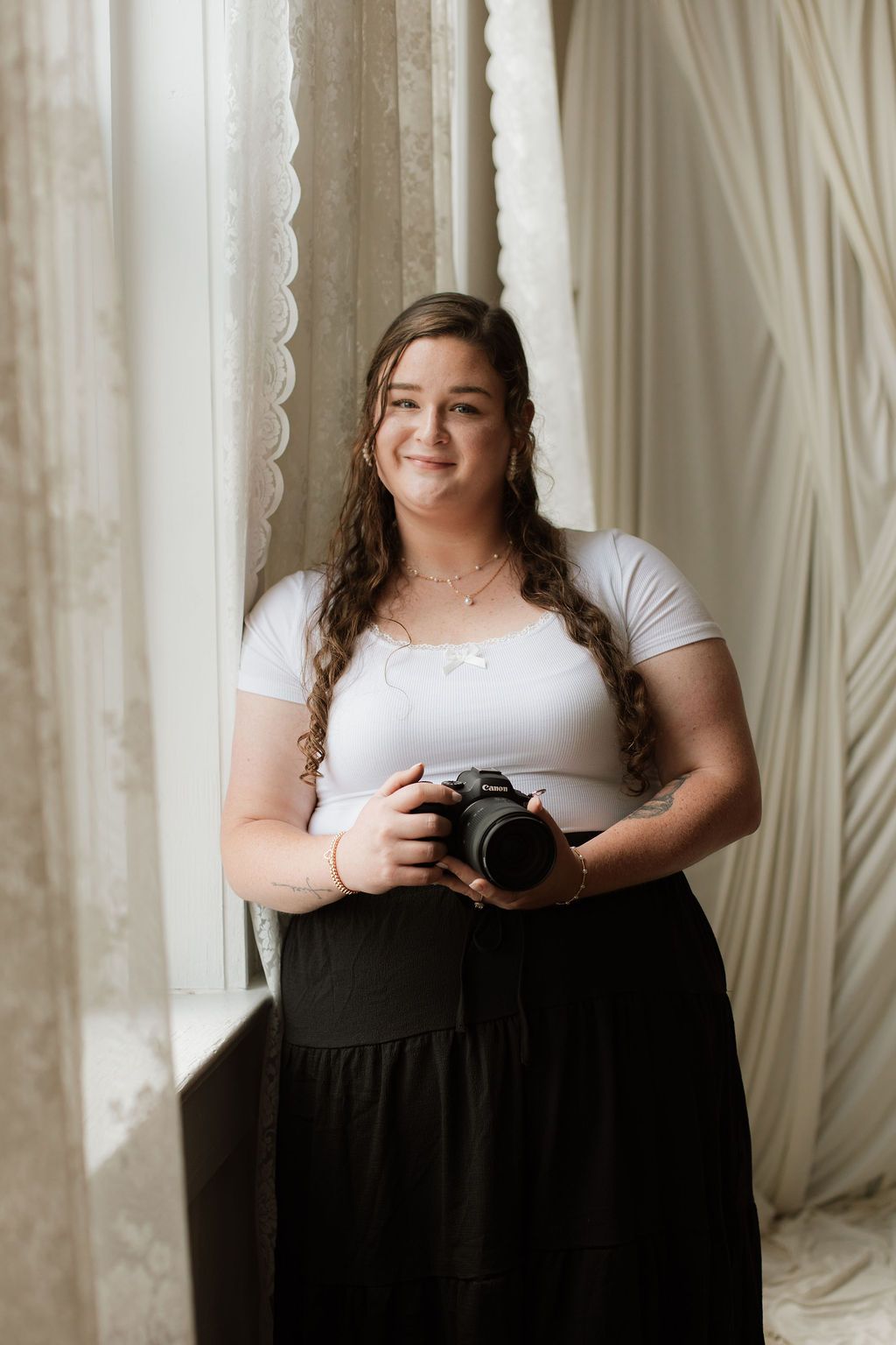Young woman with curly hair holding a DSLR camera, wearing white top and black skirt, standing by a window.