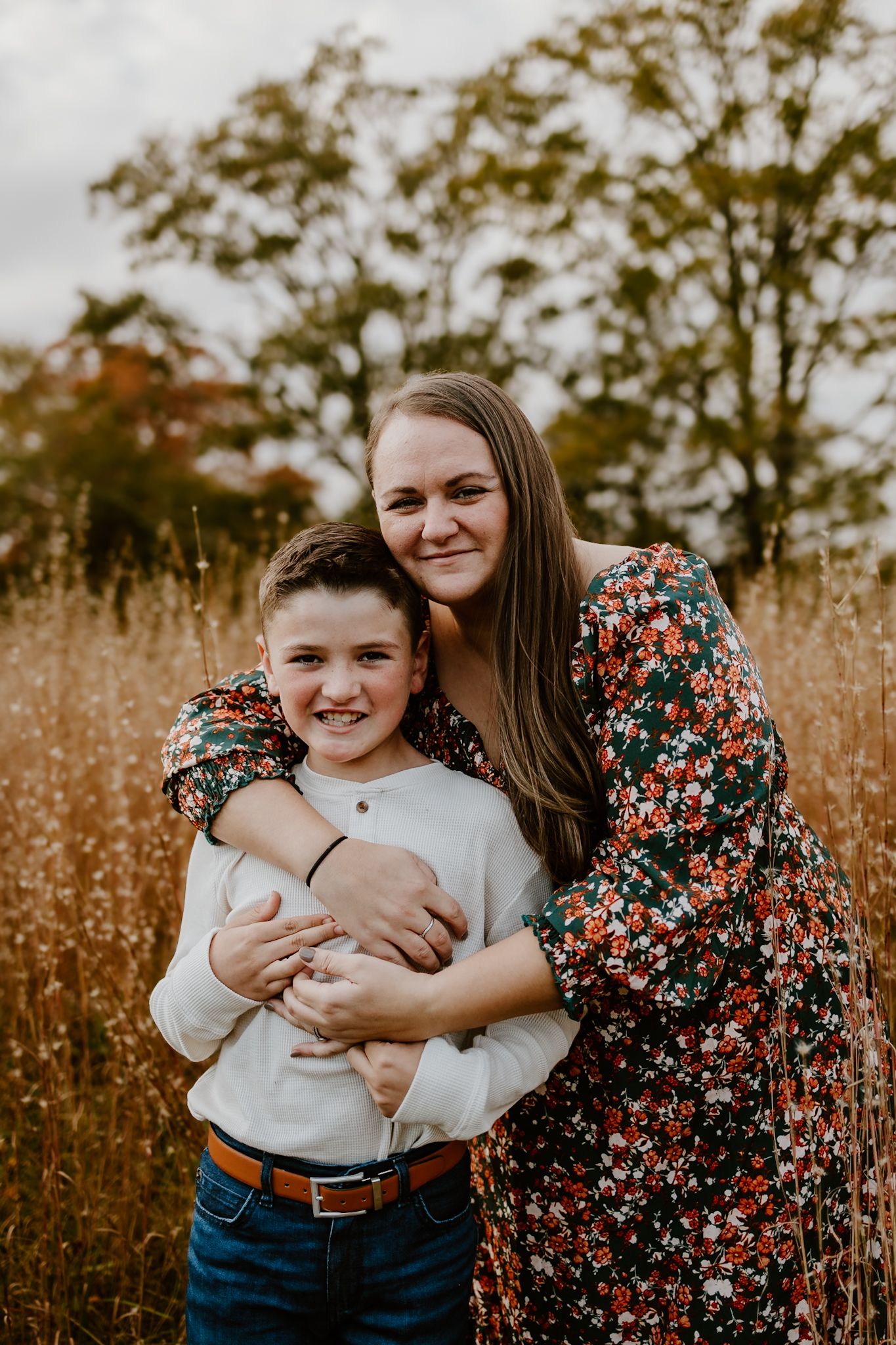 A woman in a floral dress hugs a smiling boy in a white shirt in a golden field with autumn trees.