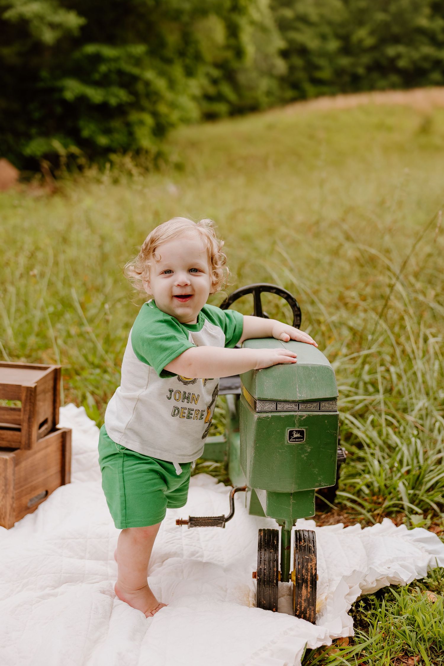 Toddler in John Deere outfit smiling while playing with a vintage green toy tractor outdoors on a white blanket.