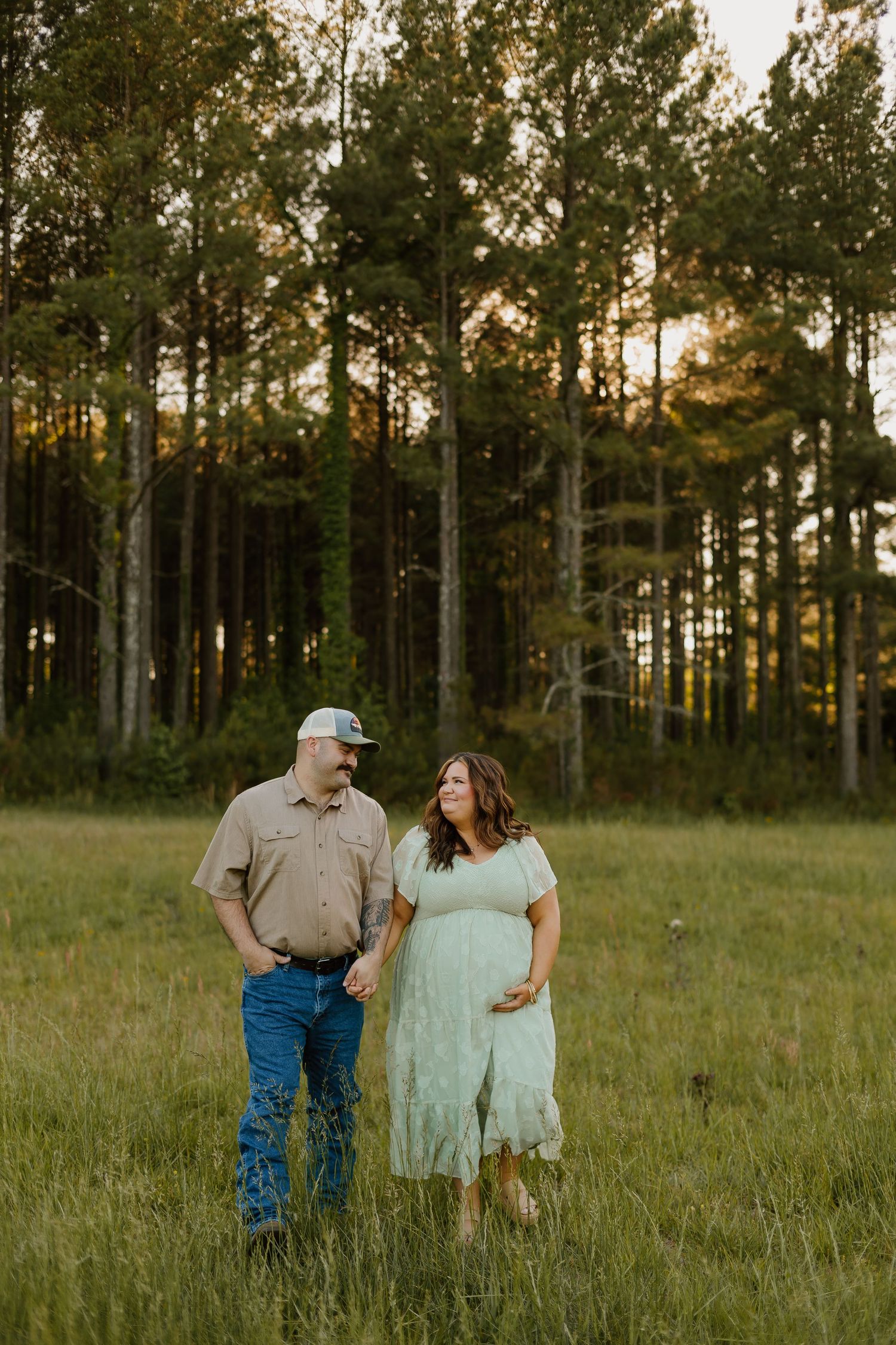 Pregnant couple holding hands in a sunlit green meadow with tall pine trees at golden hour.