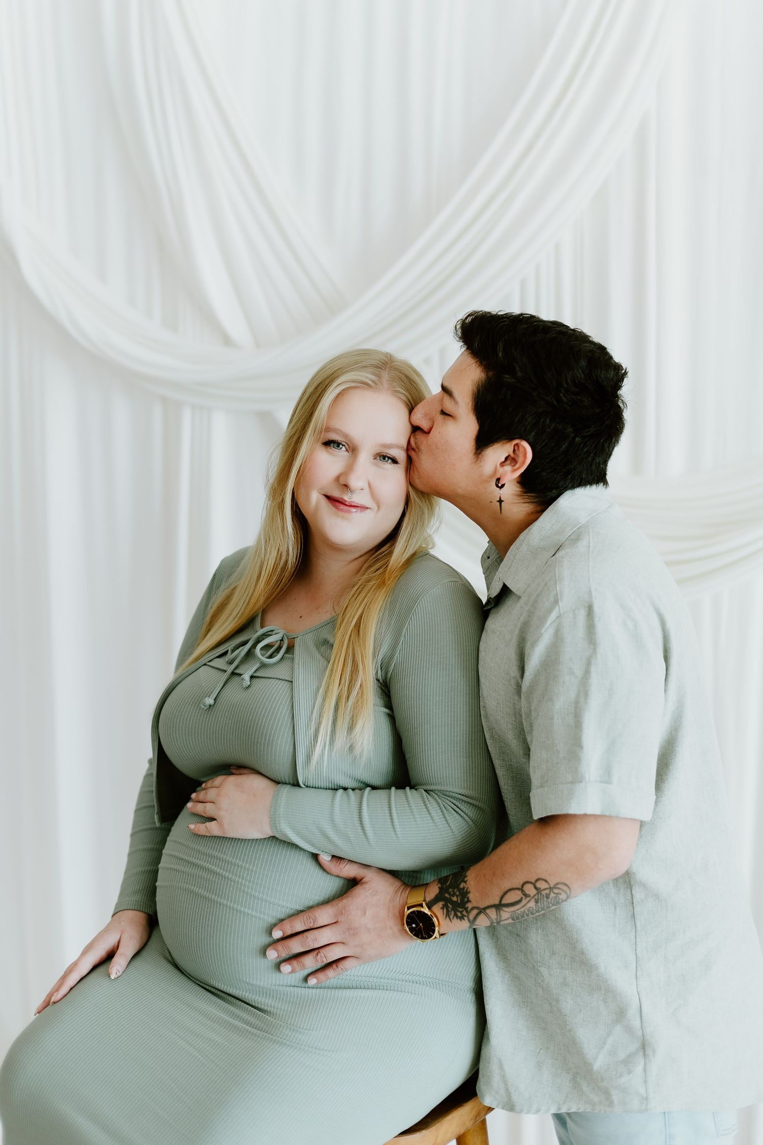 Partner kisses pregnant woman's cheek as she smiles in sage green dress in white studio setting.